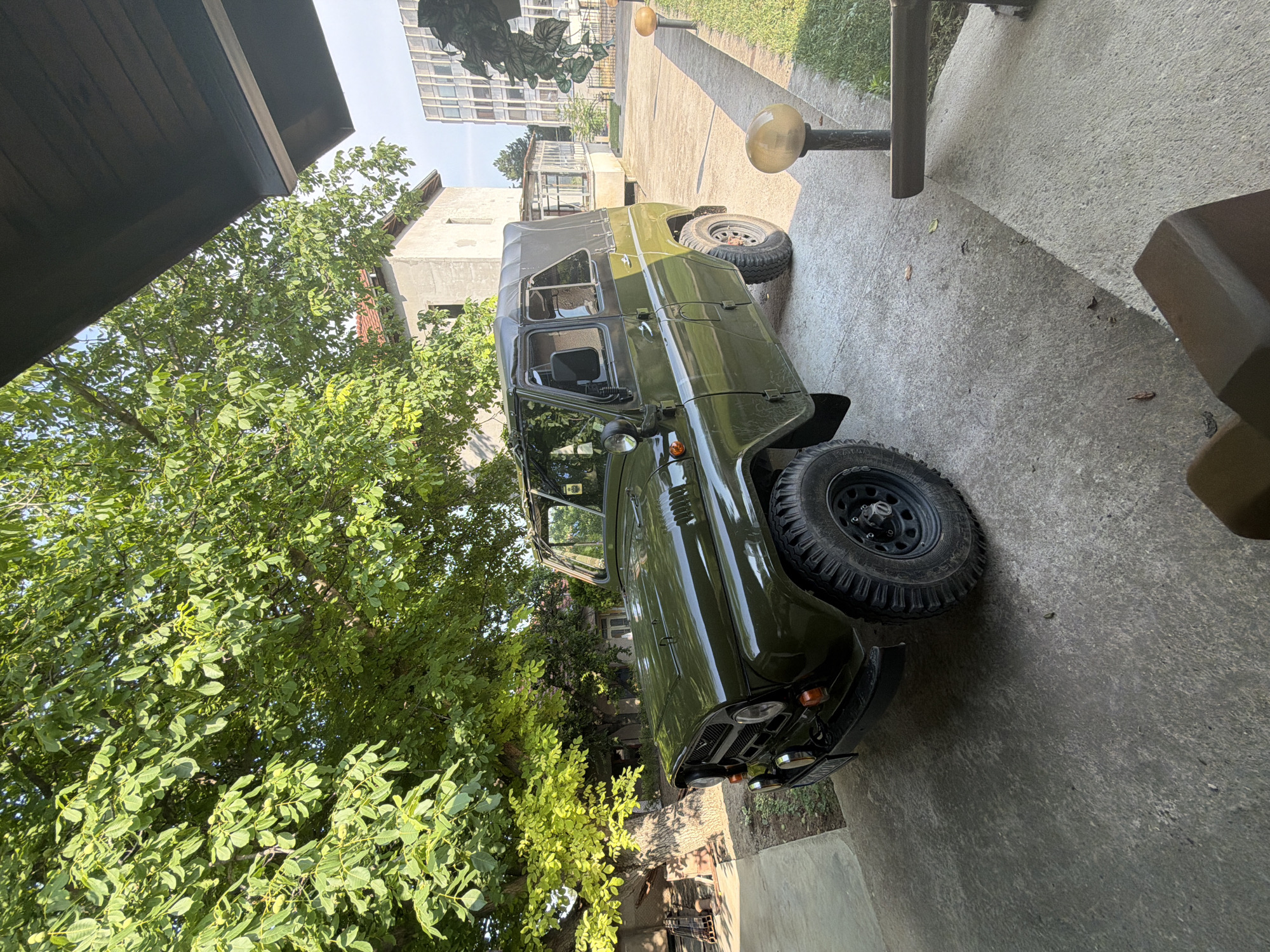 This media file depicts an outdoor scene in Knezha, Bulgaria, featuring a green UAZ-469 off-road vehicle. The vehicle is parked on a concrete surface, possibly a driveway or yard, adjacent to lush green trees and some residential buildings in the background. The UAZ is a four-door model with a soft top and appears to be well-maintained, with prominent off-road tires.
The scene is bathed in sunlight, suggesting it is daytime, likely late morning or afternoon, with clear weather conditions. Shadows are cast on the concrete, indicating the sun's position. The presence of trees with dense foliage and the style of the buildings suggest a residential or semi-rural setting.
No people are visible in the frame. The primary object of interest is the UAZ-469. The overall mood conveyed is one of stillness and perhaps a sense of ruggedness associated with the vehicle. The orientation of the image is rotated, with the vehicle appearing to be on its side relative to a standard upright view. No visible text is discernible in the image.