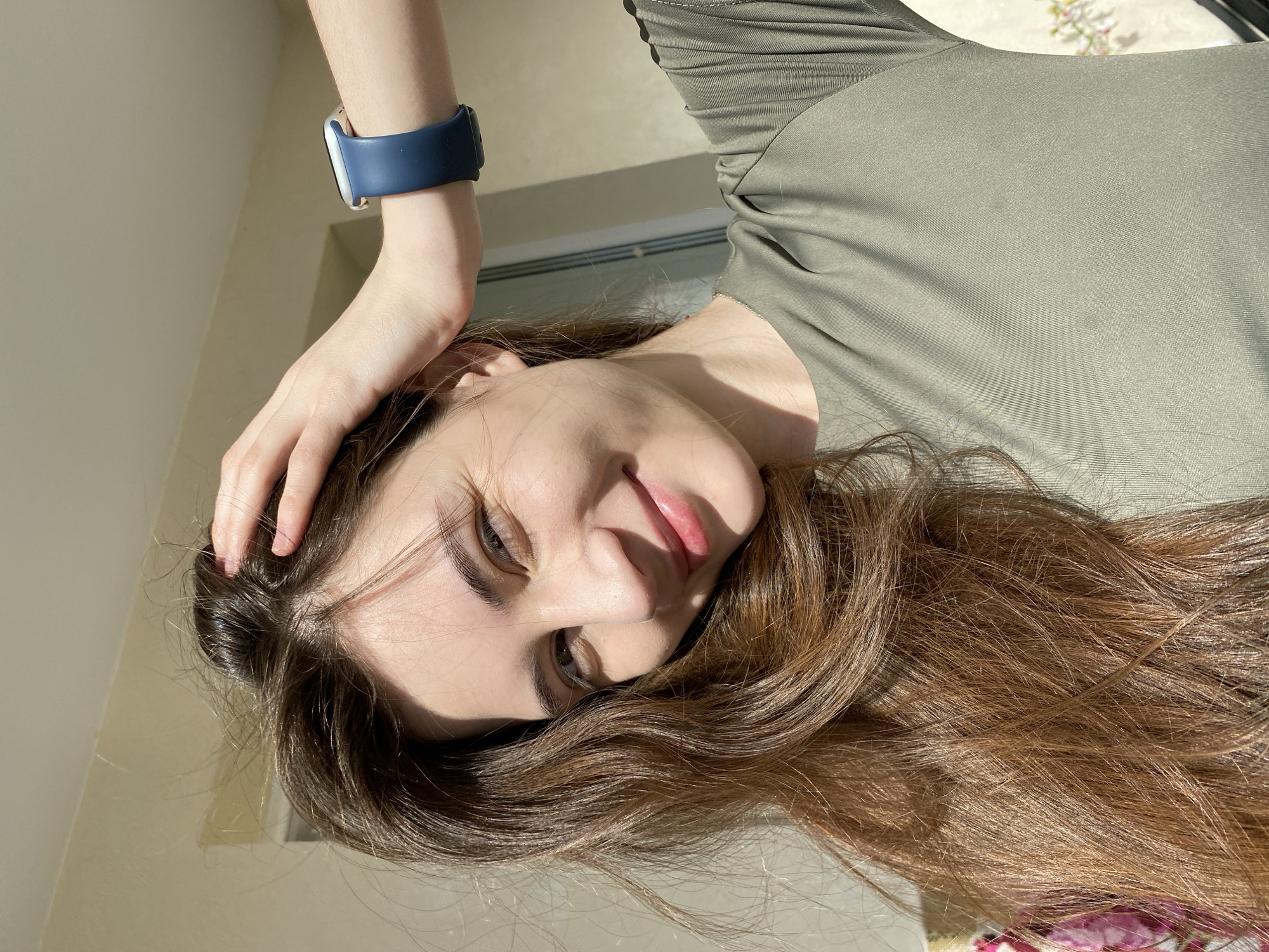 A medium close-up, portrait-style photograph depicts a young woman in an indoor setting, with the image rotated 90 degrees counter-clockwise. She is shown in a reclined or lounging pose, looking upwards towards the camera with her head tilted to her right. She has long, wavy, reddish-brown hair that flows around her head. Her expression is calm and pleasant, with a subtle, closed-mouth smile and her eyes appearing dark, looking slightly to her left. Her lips are slightly parted, revealing a soft pink hue.
She is wearing a form-fitting, long-sleeved top in an olive green or khaki color. On her left wrist, a smartwatch with a silver-colored casing and a dark blue band is prominently displayed. Her left hand is resting on her head, with fingers gently touching her hair.
The setting is an interior space, characterized by bright, natural daylight streaming in, casting distinct shadows on her face and hair, indicating it is daytime. The background features a plain, light-colored wall, possibly beige or off-white, with a hint of a window frame or blinds in the upper portion of the image. A small section of patterned fabric, potentially floral, is visible in the bottom right corner of the frame. The overall impression is one of casual relaxation under natural illumination. There is no visible text in the image.