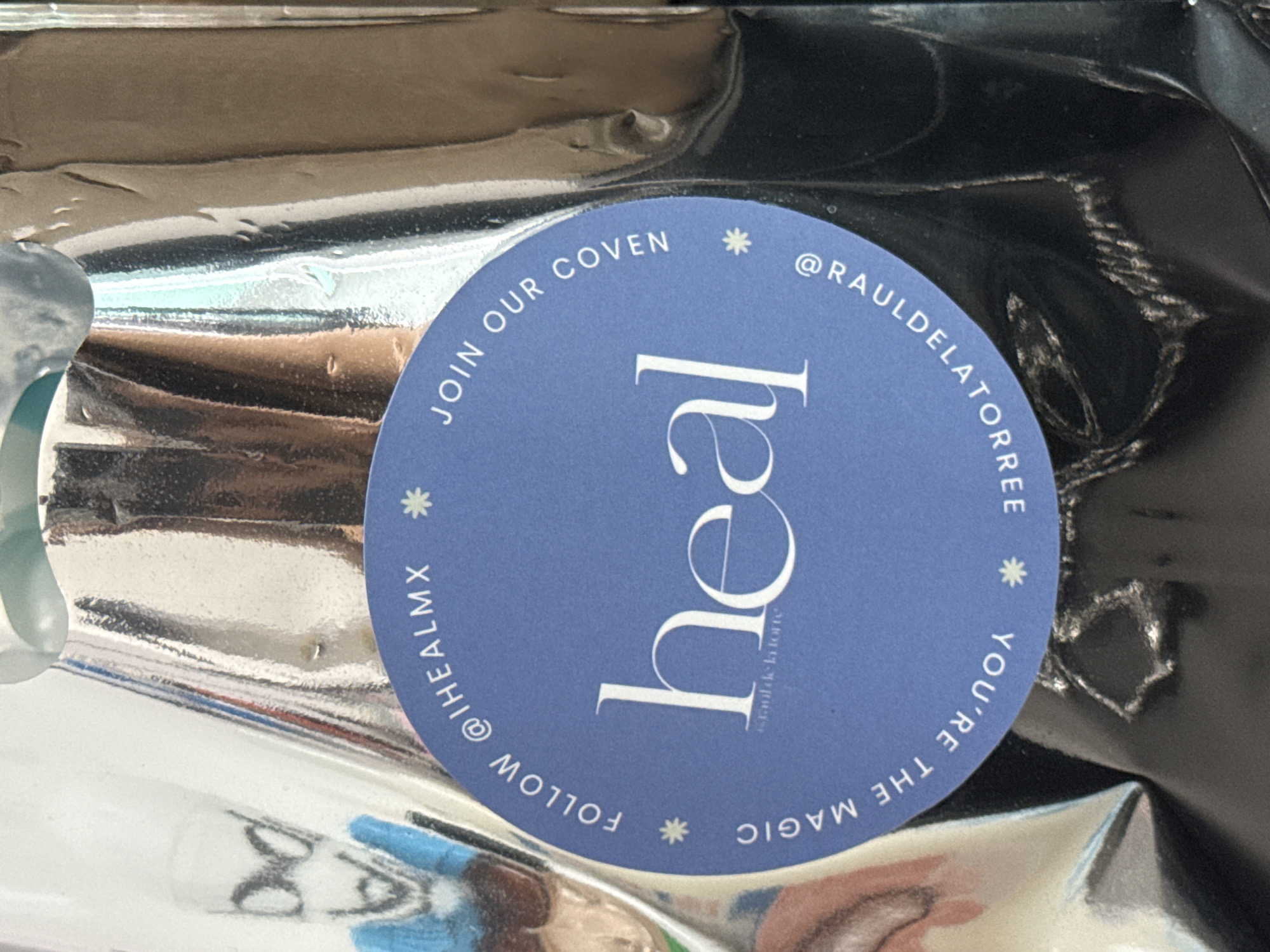 The image shows a close-up of a round, blue sticker adhered to a shiny, reflective surface, possibly a metallic package or container. The sticker has the word "heal" in a large, stylized font, with smaller text "rauldela torre" underneath. Around the perimeter of the sticker are the phrases "JOIN OUR COVEN," "@RAULDELATORREE," "YOU'RE THE MAGIC," and "FOLLOW @THEALMX," separated by asterisks. The reflective surface behind the sticker is distorted and shows fragmented reflections of other objects, suggesting an uneven or crumpled material. The location, San Bernardino Tlaxcalancingo, Mexico, is not visually represented in the image.