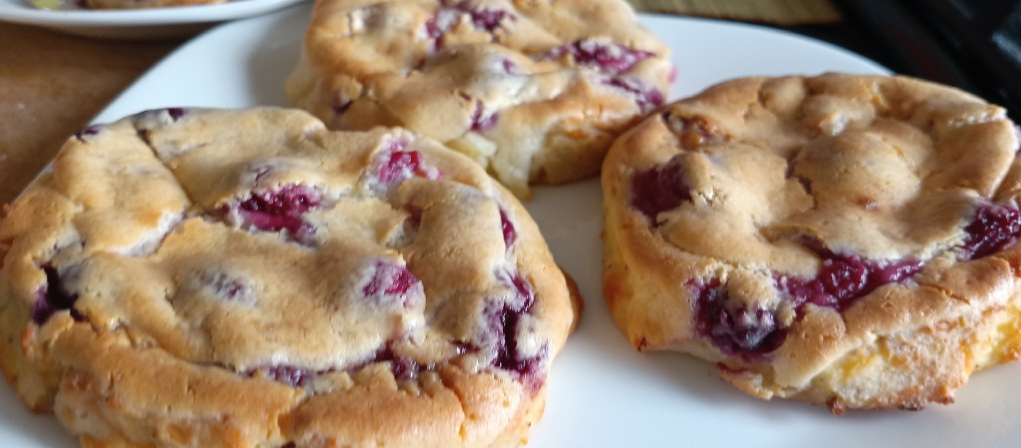 The image displays three freshly baked items, resembling muffins or small cakes, arranged on a white plate. Each treat features a golden-brown, somewhat irregular and domed top, with visible dark red or purple berries, likely raspberries or cherries, baked into the surface and sides. The texture of the baked goods appears light and fluffy, with some cracks on the surface and lighter dough showing through in places. The vibrant color of the berries creates a striking contrast against the warm, golden hue of the pastry.
The baked items are the central focus, presented neatly on a clean, white ceramic plate. In the background, a portion of another white plate is visible, suggesting more food beyond the frame, and a blurred, textured background element, possibly a wooden surface or placemat. The scene conveys an indoor setting, likely a home kitchen or dining area, where a batch of homemade treats has just been prepared. No specific activity is captured, but the presentation implies they are ready to be eaten. There are no people visible, nor any discernible text. The lighting appears soft and natural, highlighting the appealing textures and colors of the food.