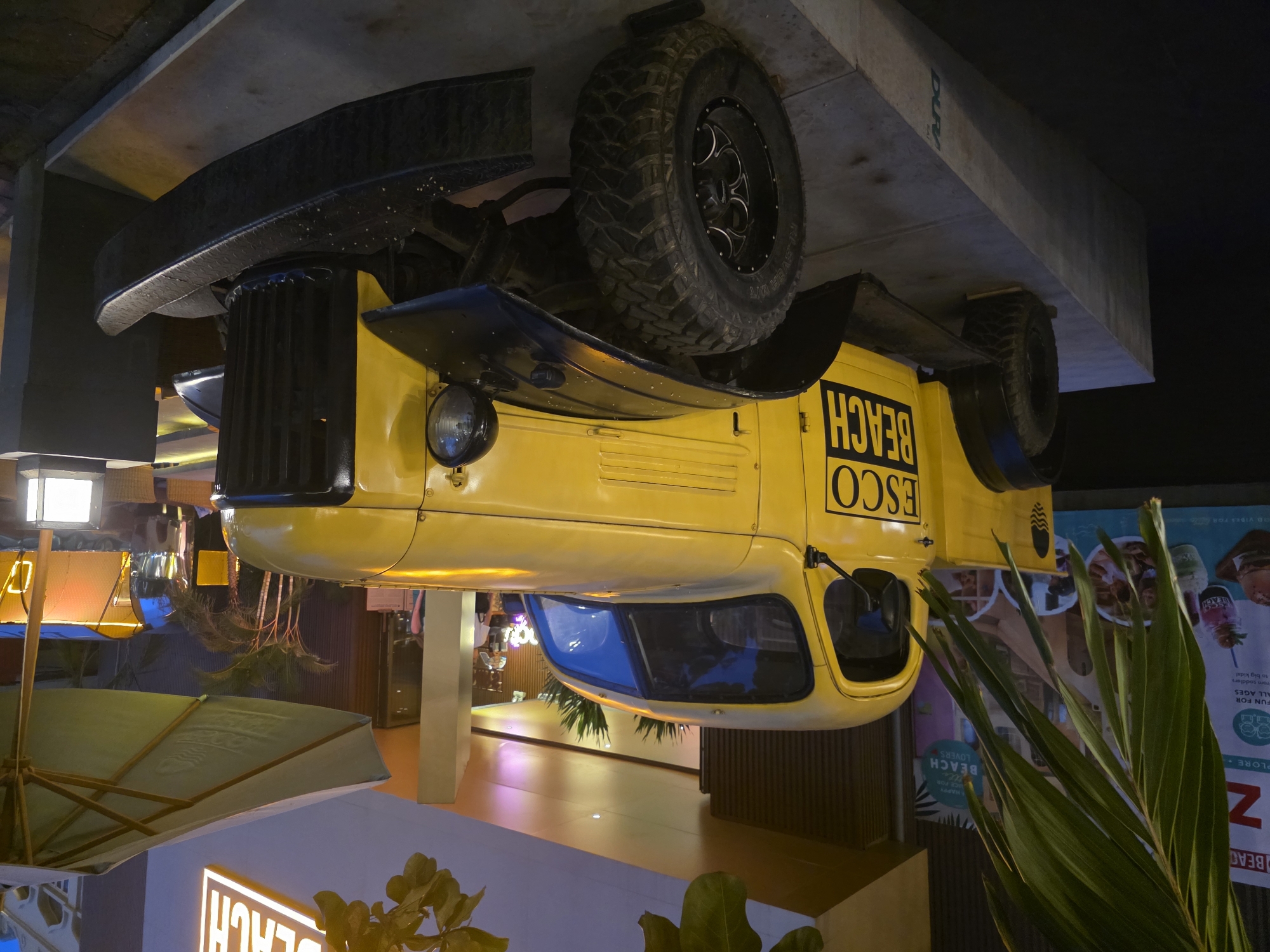 The image shows a vintage yellow pickup truck, likely a Dodge Power Wagon, parked outdoors at night. The truck has a black grille and bumper, and large off-road tires. The text "ESCO BEACH" is visible in black lettering on the side of the truck. The truck is parked on a raised concrete platform. In the background, there is a building with a lit sign that reads "BEACH" in large, white, illuminated letters. There are also palm trees and outdoor seating with umbrellas visible. The scene appears to be in a commercial or entertainment area, possibly a beachfront establishment. The truck is illuminated by artificial lights, creating a warm glow. The lighting suggests it is either dusk or night. The overall impression is one of a decorative display or a vintage vehicle for rent or promotion. The presence of the "BEACH" signage and palm trees suggests a coastal location. Given the text on the truck and the prominent "BEACH" sign, this scene is likely at Esco Beach, a popular nightlife and dining spot in Đà Nẵng, Vietnam.