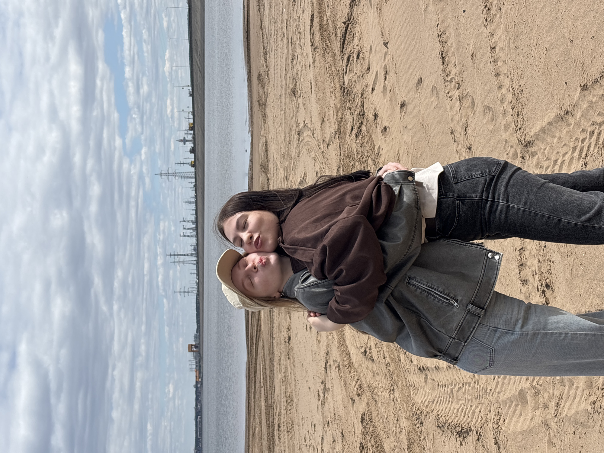 Two young women are embracing on a sandy beach. One woman has long, dark hair and wears a brown sweatshirt and dark jeans. The other has blonde hair, a beige cap, and wears a grey jacket and jeans. In the background is a body of water with a cityscape on the opposite shore, with industrial structures and power lines visible. The sky is cloudy. The scene is identified as being located near Noviy, Russia, based on the presence of specific industrial and power line features visible in the background.