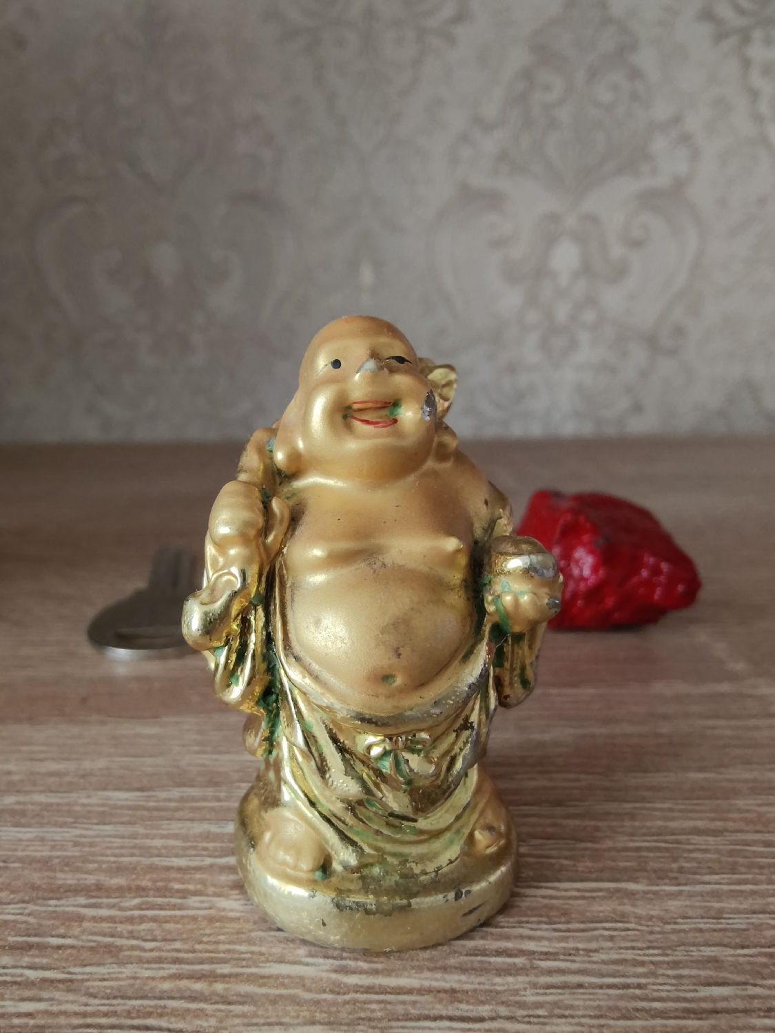 A small, golden figurine resembling a laughing Buddha (Budai or Hotei) stands centrally on a light brown, wooden surface, likely a table or shelf. The figurine has a jovial expression with a wide smile, red lips, and black dot eyes. Its golden paint shows signs of wear, particularly on the nose, chin, chest, hands, and base, revealing a lighter material underneath. Green accents are visible around its mouth and in the folds of its robes. It appears to be holding small, indistinct objects in its hands, with a rounded object visible in its right hand.
To the left of the figurine, a metallic object, possibly a key or part of a keychain, is partially visible and blurred. To its right, a red, irregularly shaped object with a textured surface, possibly a decorative stone or art piece, is also blurred.
The background consists of a light-colored wall adorned with a subtle, embossed or patterned wallpaper featuring an ornate, scroll-like design, which is out of focus. The scene is illuminated by soft, even lighting, suggesting an indoor setting during the daytime. No specific activity is depicted other than the arrangement of these objects. There is no visible text in the image. The location context for this scene is Kryvyi Rih, Ukraine.