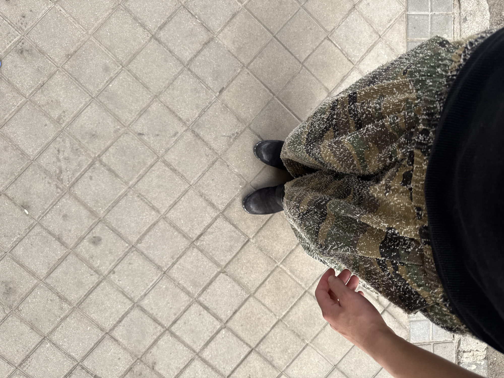 The image is a high-angle, top-down shot capturing the lower body of a person standing on a paved outdoor surface. The person wears dark, possibly black, boots and a voluminous garment, likely a long skirt or wide pants, featuring a camouflage pattern in shades of green, brown, and black. This garment has a distinctive textured appearance, flecked with numerous small, white or light-colored fibers or dots, giving it a somewhat fuzzy or frosted look. A black garment, possibly a sweater or top, is visible on the far right, partially obscuring the person's upper body.
A light-skinned hand extends from the lower right of the frame, gently touching or resting near the camouflage garment. The ground is paved with light grey, square tiles arranged in a diagonal grid pattern, typical of an urban sidewalk or public space. The tiles show some signs of wear and minor dirt accumulation. The even, bright lighting suggests it is daytime. The person appears to be standing still. No text is visible in the image. The paving style is consistent with urban environments like Madrid, Spain, as indicated by the provided context.