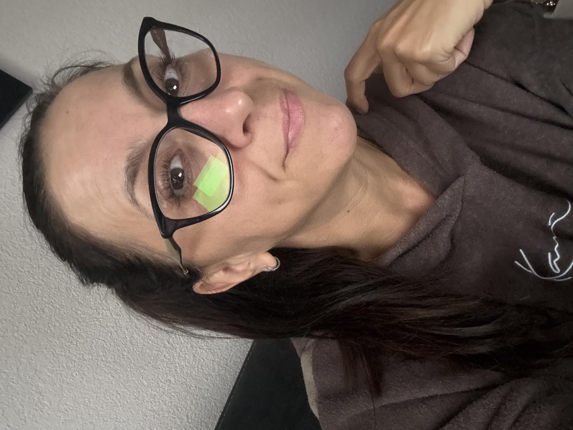 A woman with dark hair pulled into a ponytail and wearing black-rimmed glasses is looking at the camera. She is wearing a brown hoodie with a white graphic logo. The background is a textured grey wall. She has a small silver hoop earring in her left ear. Her right hand is resting on her shoulder. Two green rectangular shapes are reflected in her left lens. The image was taken in Marum, Netherlands.