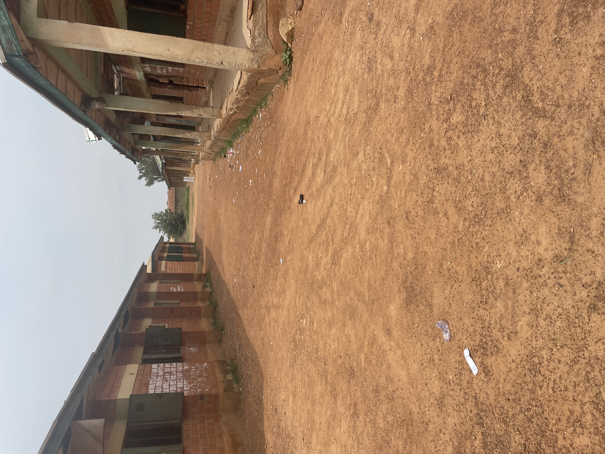 The image depicts an outdoor scene in Television, Nigeria, showing a pathway between two brick buildings. The pathway is covered in reddish-brown dirt, with scattered debris, including pieces of paper and a dark object that resembles a shoe or a piece of footwear.
The buildings appear to be school classrooms or similar structures. The building on the left is made of red brick with green-shuttered windows and shows some faded writing on its exterior walls. The building on the right has a covered walkway supported by concrete pillars, also constructed with red brick, and has visible signs of wear and tear. The sky above is overcast, suggesting a cloudy or hazy day. The overall environment appears to be a school compound or a similar institutional setting, with an unpaved ground surface and basic architectural structures.