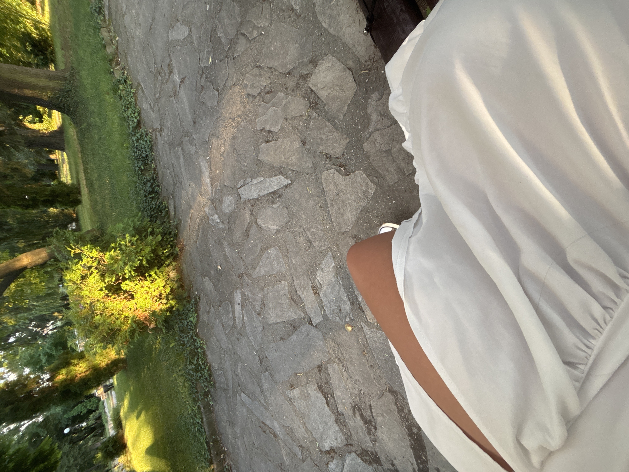 The image shows a person sitting on a bench in an outdoor setting, likely a park or garden. The foreground is dominated by the person's white clothing, which is draped over their lap and legs. A glimpse of the person's legs and a white sandal are visible. The background reveals a stone pathway, a grassy area with ivy, and several trees. The sunlight casts shadows and highlights on the scene. The location is identified as the town of Garmen, Bulgaria. There are no other individuals visible in the image, and the focus is on the person's attire and immediate surroundings. The scene is quiet and appears to be captured during daylight.