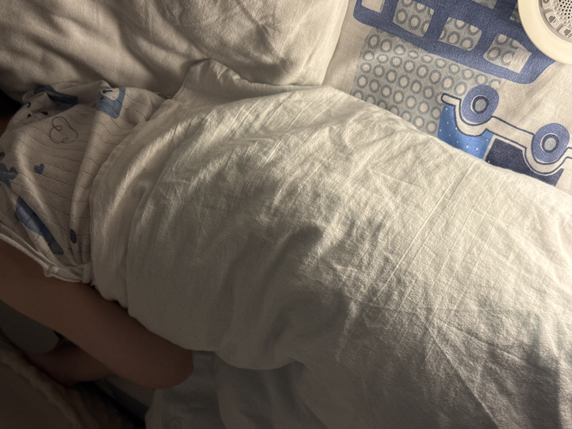 The image shows a close-up, overhead view of a young child sleeping in a bed, covered by a white duvet. The child's arm is visible on the left side, and they are wearing pajamas with a blue and white pattern featuring cartoonish animals and hearts. The bedsheets also have a blue and white pattern with cartoon buses. The scene appears to be a bedroom in a residential setting, likely in Lyubertsy, Russia. The lighting suggests it is either early morning or evening, as there are shadows cast on the duvet.