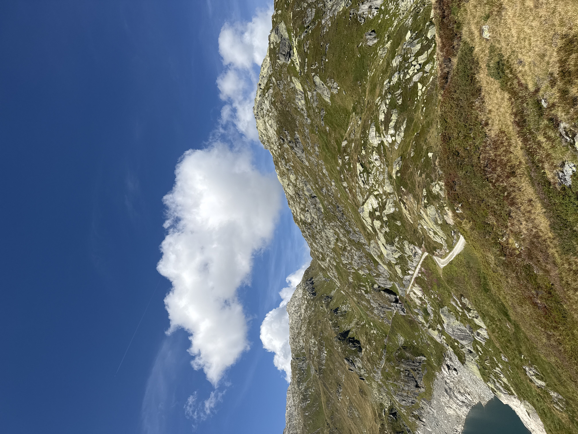 The image depicts a scenic mountain landscape under a clear blue sky with scattered white clouds. A winding dirt road traverses the grassy, rocky slopes of the mountains. In the foreground, dry, golden grasses and low-lying shrubs are visible. To the left, a dark blue body of water is partially visible at the base of a steep, rocky incline. The mountains themselves are covered in patches of green grass and large, light-colored rocks. A contrail from an aircraft is faintly visible in the upper left portion of the sky. The scene is characteristic of the alpine environment found in the vicinity of Airolo, Switzerland, likely at a higher elevation. There are no people or visible man-made structures apart from the road.