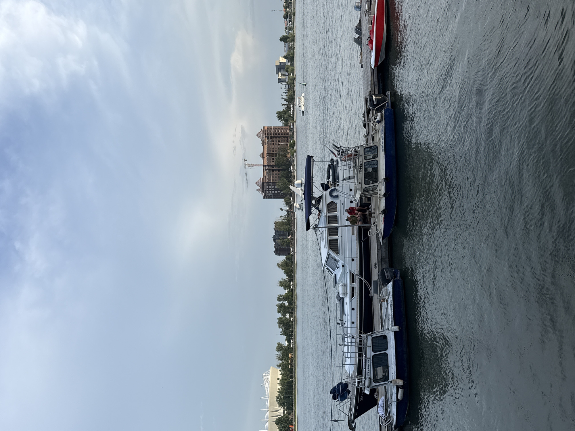 The image shows a view of Rostov-on-Don, Russia, taken from a waterfront location. Several boats are docked parallel to the edge, the closest of which is a white and blue vessel with visible script in Cyrillic. A red boat is seen moored alongside the white one. Across the water, a cityscape is visible featuring lush trees, tall buildings, and what appears to be a construction crane near an ornate palace or castle-like structure. The sky is partly cloudy.