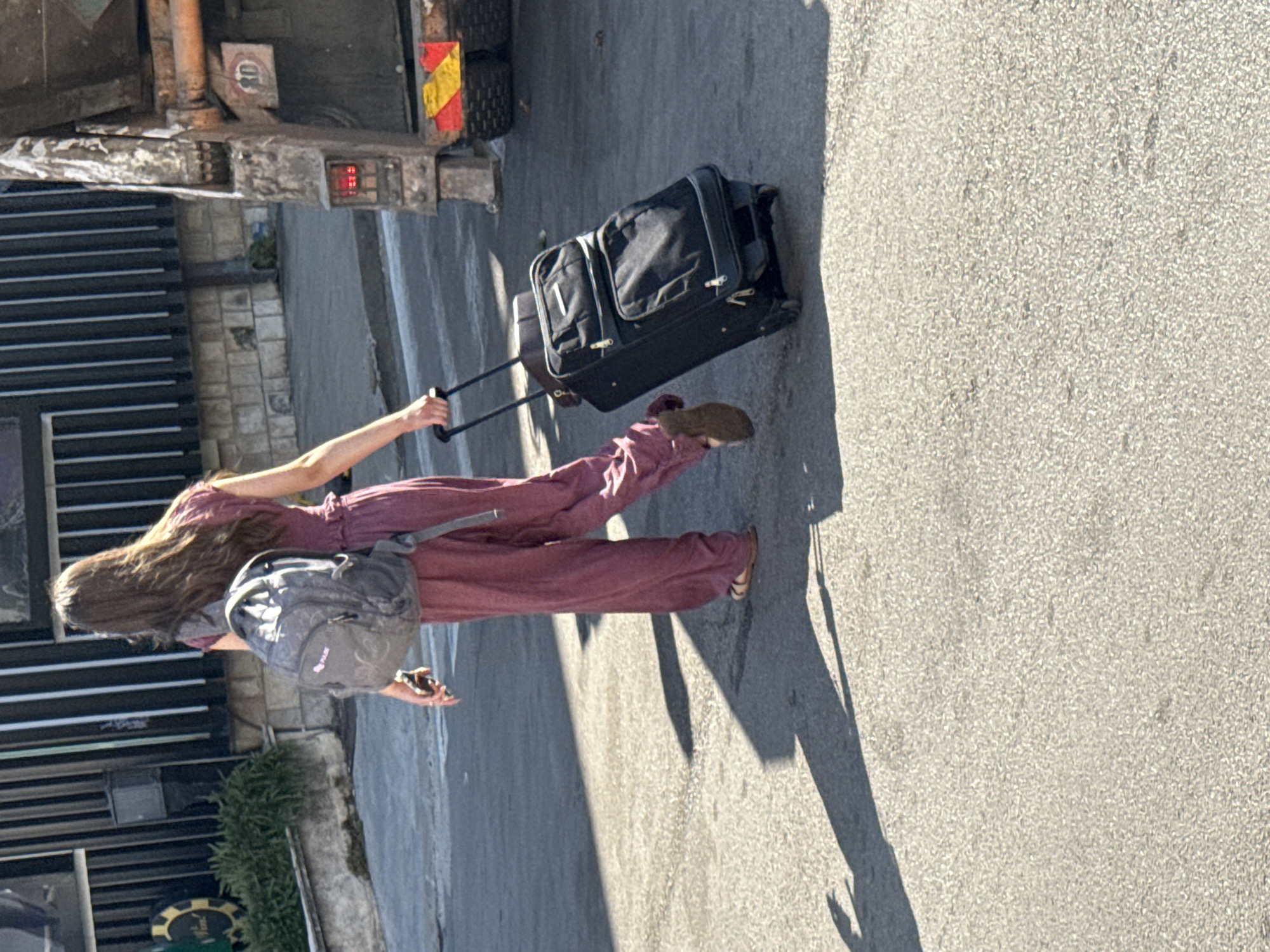 A young woman with long, dark hair, wearing a maroon jumpsuit and brown sandals, walks away from the camera on a paved street in Budva, Montenegro. She is carrying a gray backpack on her shoulders and pulling a black rolling suitcase. The suitcase has a retractable handle and is being rolled alongside her. In the background, a large, weathered truck is parked, featuring reflective tape and a speed limit sign. A building with vertical slats and a stone facade is visible behind the woman. The ground is a typical asphalt street, with distinct shadows cast by the woman and her luggage, indicating a sunny day.