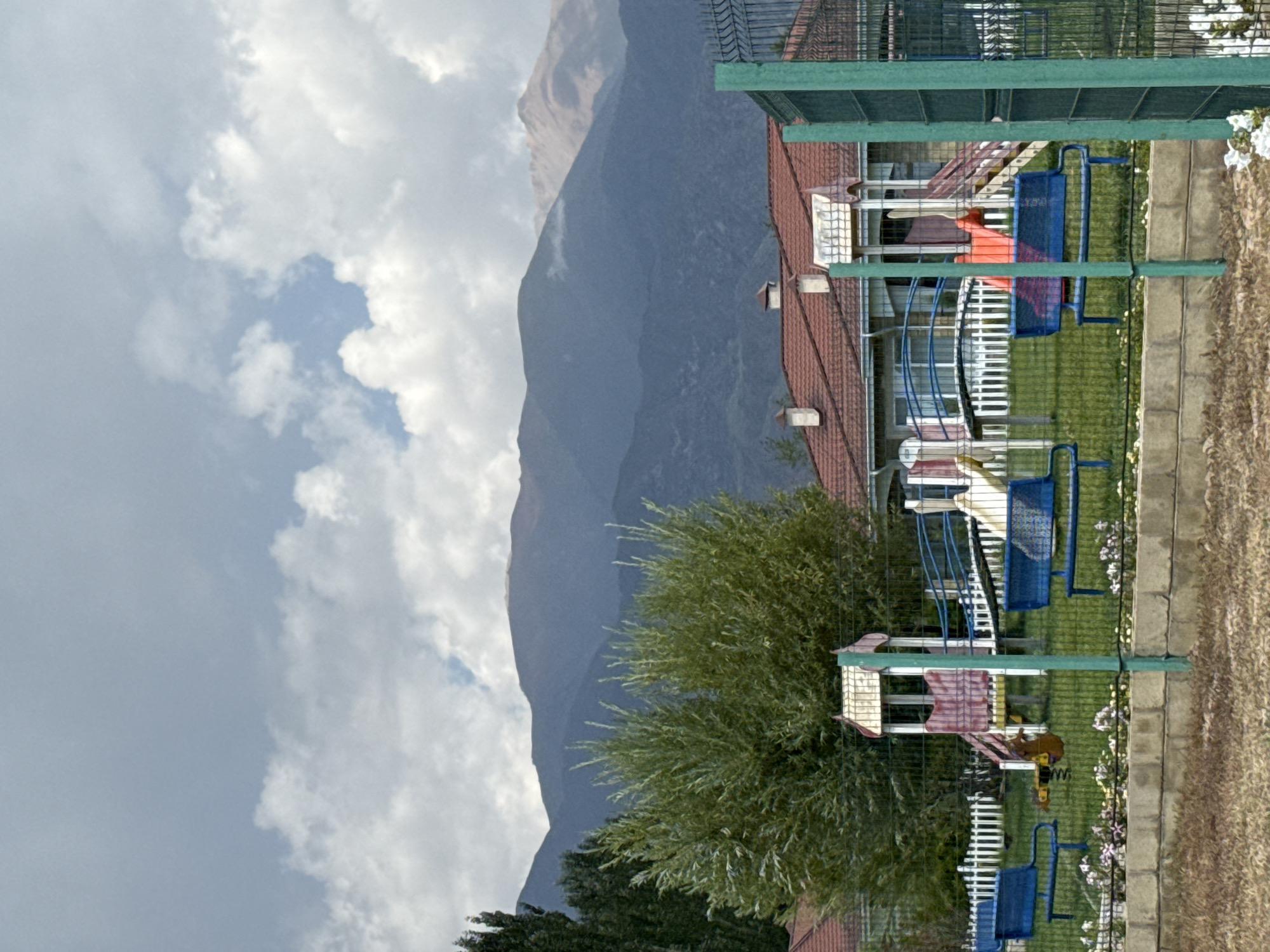 This is a medium-shot, eye-level, outdoor photograph taken in the city of Sary-Oy, Kyrgyzstan. The scene depicts a playground in front of a building, with mountains in the background.
In the foreground, a low, concrete block wall runs horizontally across the frame, with a dry, patchy brown grass ground surface in front of it. A metal fence, with a green pole in the center, divides the foreground from the midground.
The midground features a playground with several pieces of equipment. There are two blue park benches, one on the left and one on the right, facing the playground. In the center, a multi-colored play structure includes a red slide, a white tower with a red roof, and stairs. To the left of the play structure, a white picket fence borders a section of the playground. A small, brown spring rider toy is visible near the left bench. Various small, pink and white flowers are planted in a strip along the base of the concrete wall.
Behind the playground, a building with a red tile roof and white chimneys is visible. The building appears to be a house or a small resort. Trees, including a tall, dark green tree on the far left and a bushy green tree in the center, are positioned behind the playground and in front of the building.
The background is dominated by large, layered mountains. The mountains are a deep blue-gray, with some lighter patches of brown and tan on their upper slopes. The sky above is a cloudy, overcast gray.
The photograph appears to have been taken on a cloudy day, with muted lighting. The overall impression is one of a rural or semi-rural setting, possibly a holiday resort given the playground and the proximity to the mountains. No people are visible in the frame.