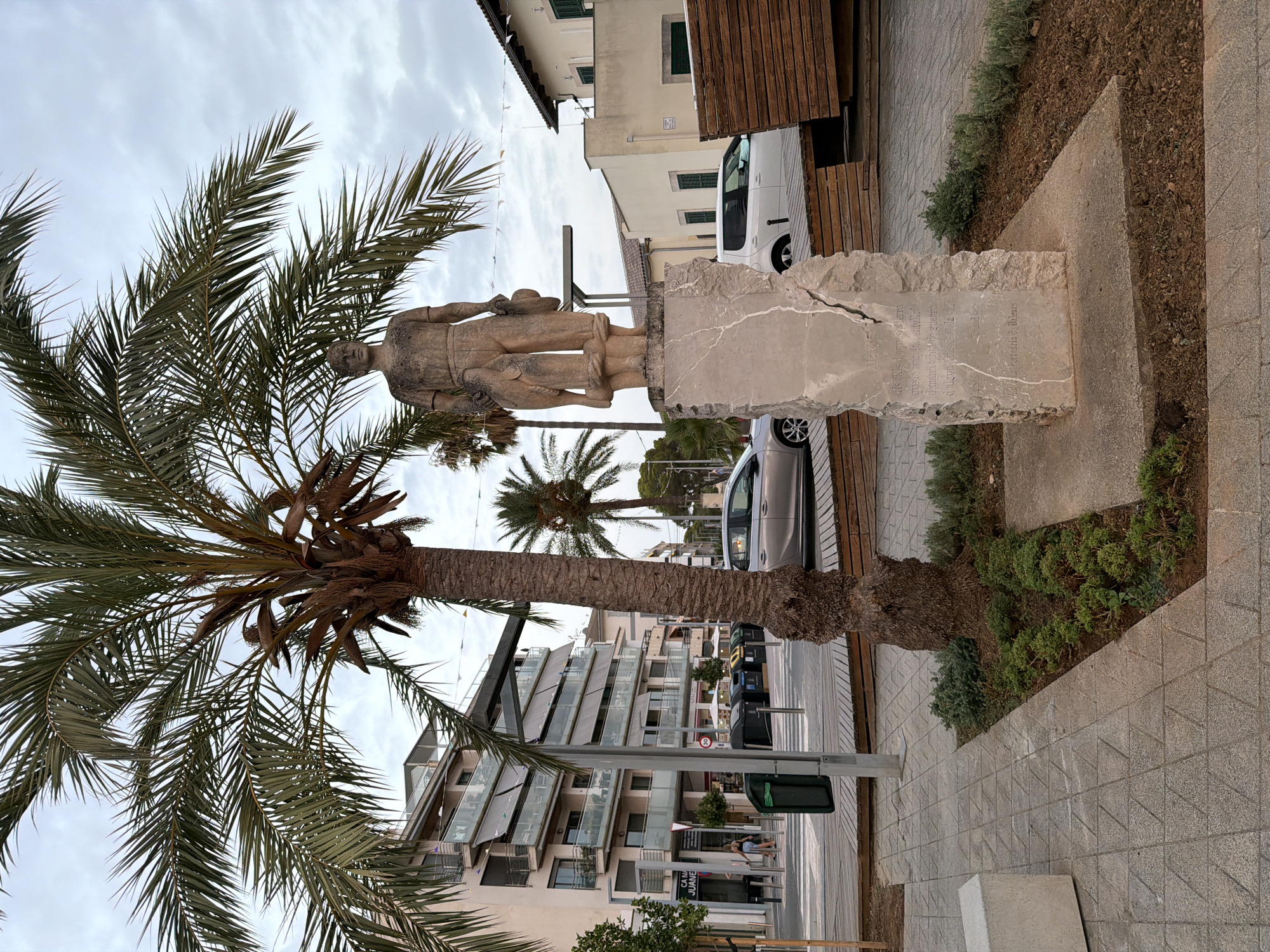 The image depicts a public square in Port d'Alcúdia, Spain, with a statue of a man and a child in the foreground. The statue, made of stone, stands on a rough-hewn stone pedestal. The man is depicted wearing simple clothing and holding a hat. Behind the statue, a large palm tree dominates the scene, its fronds casting shadows. In the background, a modern building with balconies and awnings is visible, along with other palm trees and some parked cars. The ground is paved with textured tiles, and there are some wooden benches and planters with greenery. The sky is overcast. The overall impression is that of a pleasant, somewhat tropical urban environment.