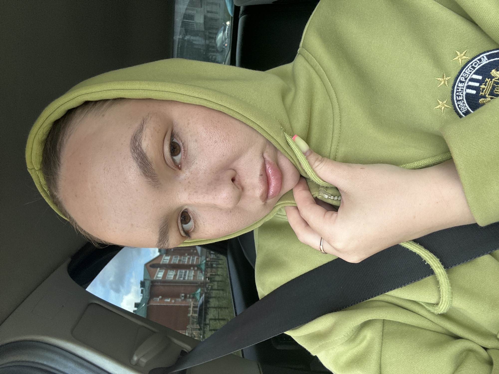 A young woman with light skin, light brown eyes, and light brown hair is sitting in the passenger seat of a car. She is wearing a light green hooded sweatshirt and a black seatbelt. Her right hand is holding the zipper of the sweatshirt. She is looking at the camera with her lips puckered. Outside the car window, there are apartment buildings and trees. The image appears to have been taken in Chaikovsky, Russia.