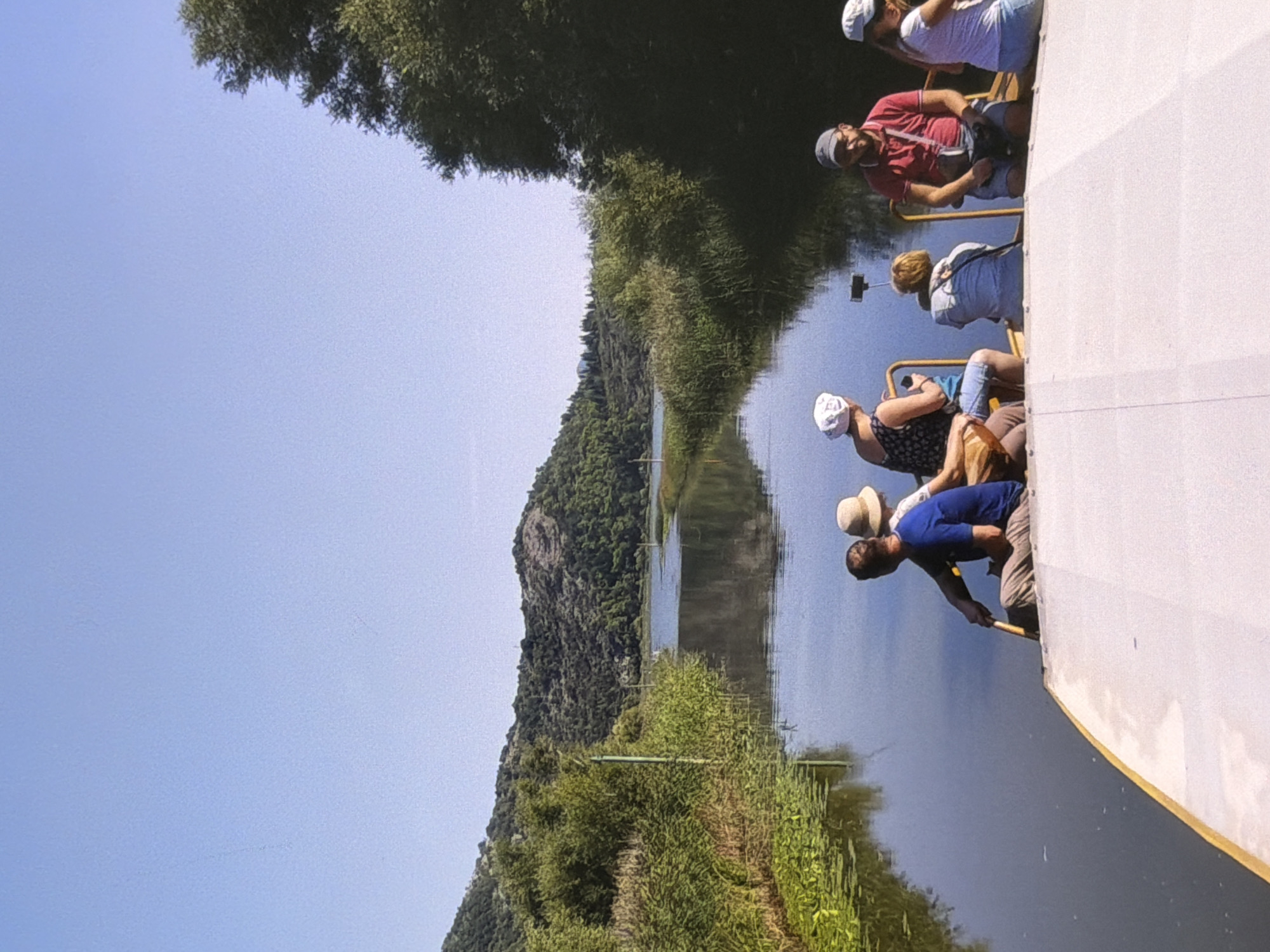 The image depicts a group of people on a boat on a calm body of water, likely a river or lake, with lush green hills in the background under a clear blue sky. The boat appears to be a flat-bottomed vessel, possibly a tour boat, with several individuals seated at the rear.
In the foreground, on the boat, are several people. A man in a blue shirt and light-colored pants is holding a paddle, suggesting he is rowing or steering. Next to him, a person wearing a straw hat is seated, possibly the person he is rowing. Further to the right, a woman in a white headscarf and patterned top is sitting, looking away from the camera. Another individual, whose back is to the camera, is wearing a light blue jacket and has short blonde hair. To the far right, a man in a red polo shirt and a baseball cap is seated, holding a camera or phone. Next to him, a woman in a white t-shirt and blue shorts is seated, also looking away. The boat's deck is a light color, possibly white or cream, with a yellow trim along the edge.
The environment surrounding the boat is dominated by water and vegetation. The water is dark and reflects the sky and surrounding greenery. Lily pads or other aquatic plants are visible on the surface of the water to the left. Tall reeds and other marshy vegetation line the banks of the waterway. In the distance, rolling hills covered in dense green trees rise up, with a lighter blue body of water visible further inland. The overall impression is one of a serene natural landscape, possibly a tourist excursion on a waterway.
