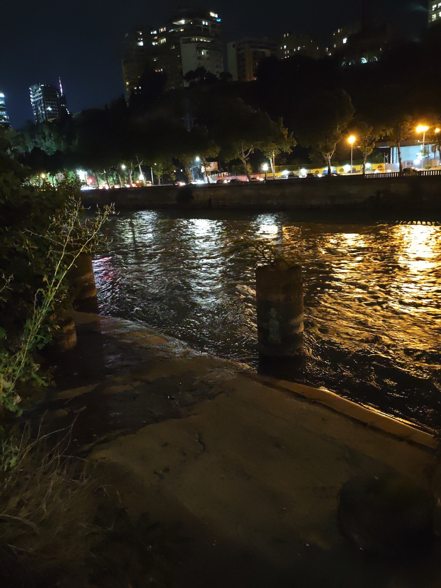The image depicts a nighttime scene along a riverbank in Tbilisi, Georgia. The river's surface is illuminated by the warm glow of streetlights and building lights, creating shimmering reflections. On the left bank, there are trees and a slope leading up to buildings. On the right bank, a walkway or embankment is visible, lined with streetlights and a railing. Beyond the embankment, several multi-story buildings are illuminated, suggesting an urban setting. The overall atmosphere is one of a city at night, with the river serving as a prominent feature of the landscape.