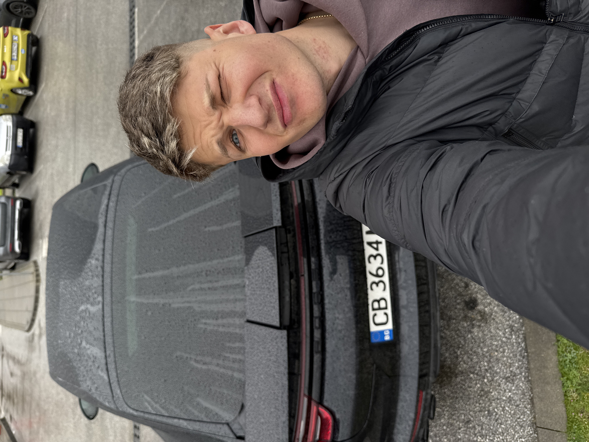 A man is taking a selfie with a black car. The car's license plate reads "CB 3634" and indicates it is registered in Bulgaria. Due to the license plate, the location can be reasonably inferred to be Plovdiv, Bulgaria. The car's surface is covered with water droplets, suggesting recent rain or washing. Several other cars are parked nearby. The ground appears to be pavement and there is a patch of grass visible. The man is wearing a dark hooded jacket and a lighter-colored sweatshirt underneath. He is winking.