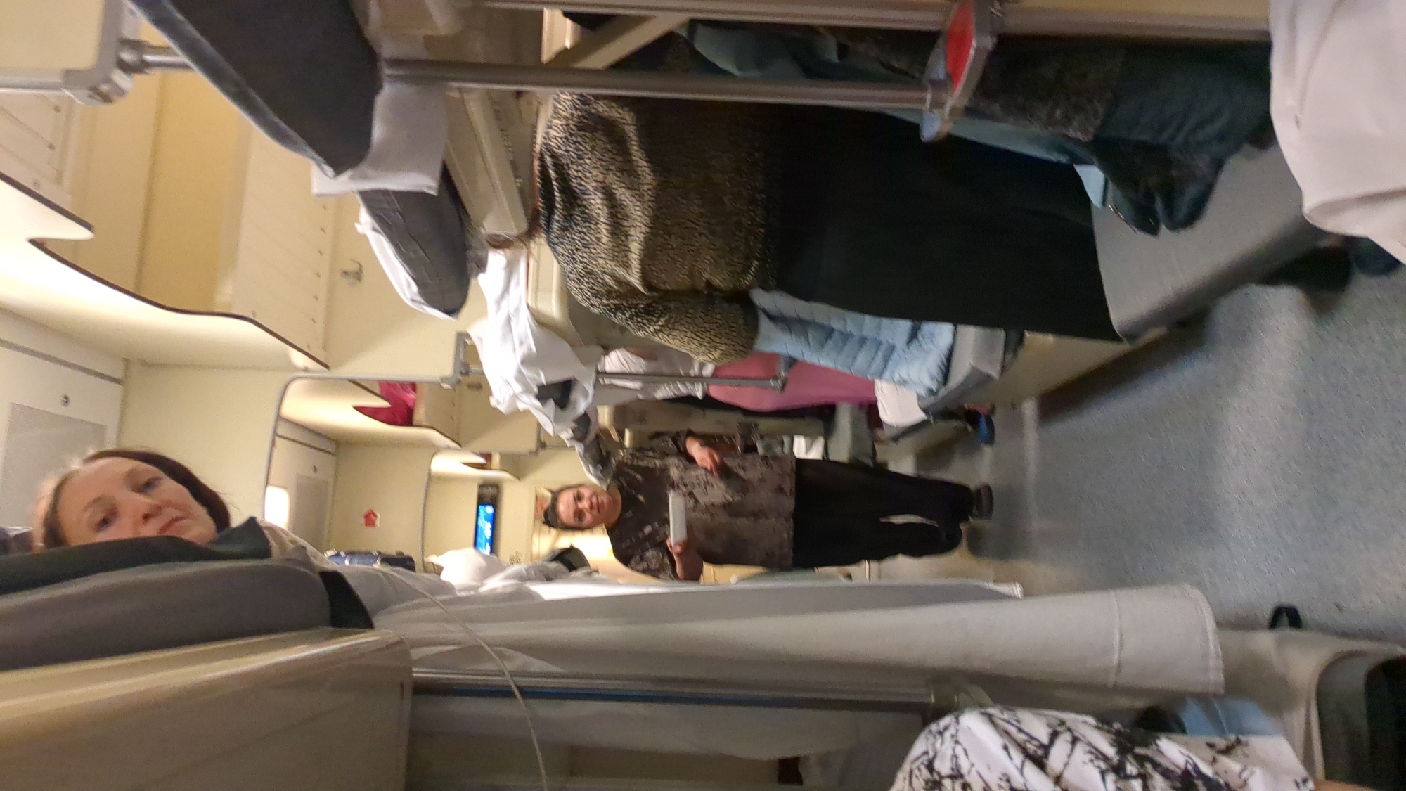 A woman is lying on a bed in what appears to be a train compartment. Another woman is standing in the aisle. Suitcases and clothes are stored on a shelf above. The interior of the train compartment is visible in the background. The location, Ulyanovsk, Russia, cannot be determined from this image alone.