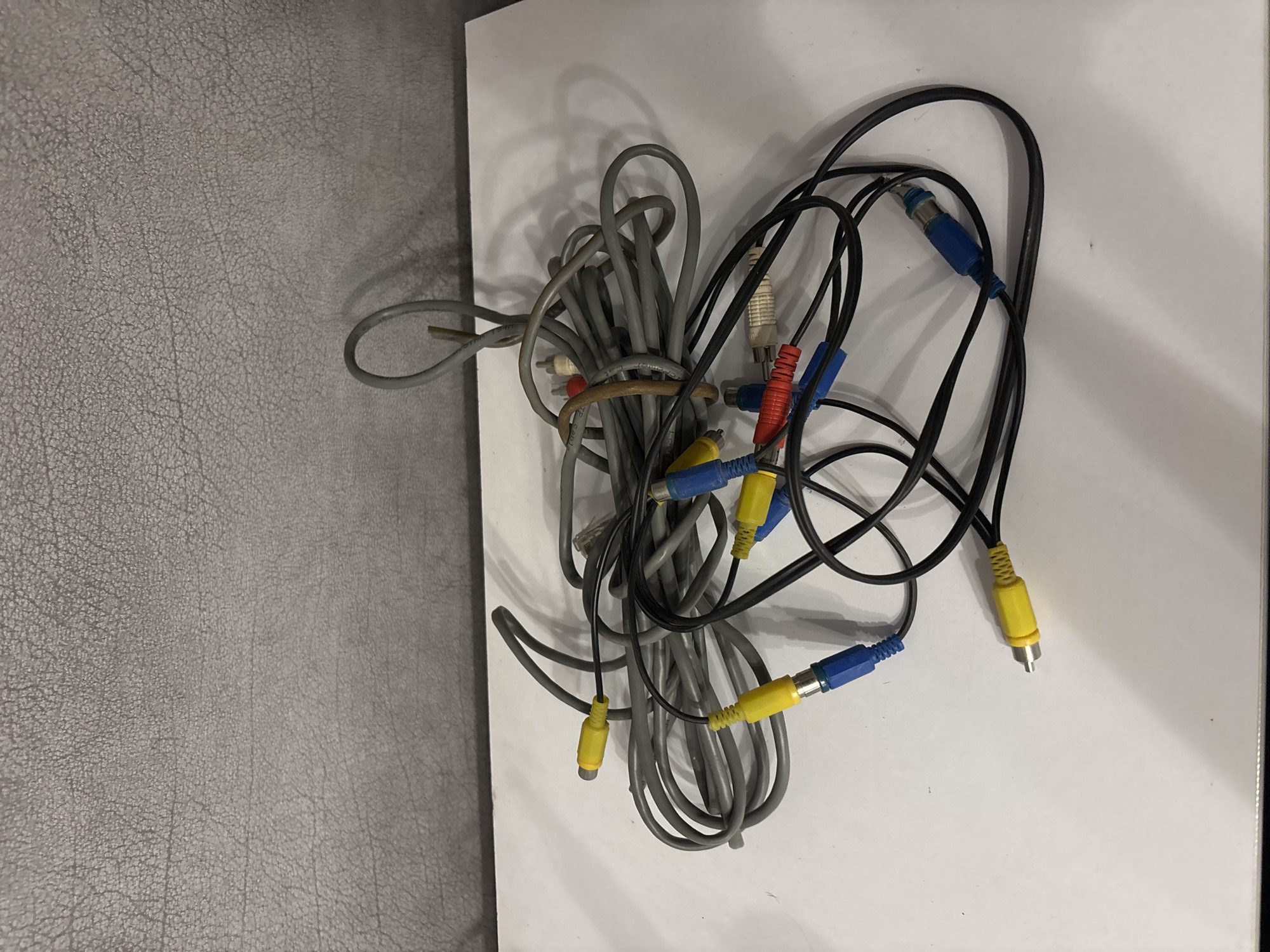 A tangled mess of audio and video cables lies on a white surface against a textured gray background. The cables are of various colors, including gray, black, yellow, red, and blue. Several RCA connectors are visible, as well as what appears to be a phone cable. The lighting casts shadows on the white surface. The scene is indoors, with no indication of a specific city or outdoor environment.
