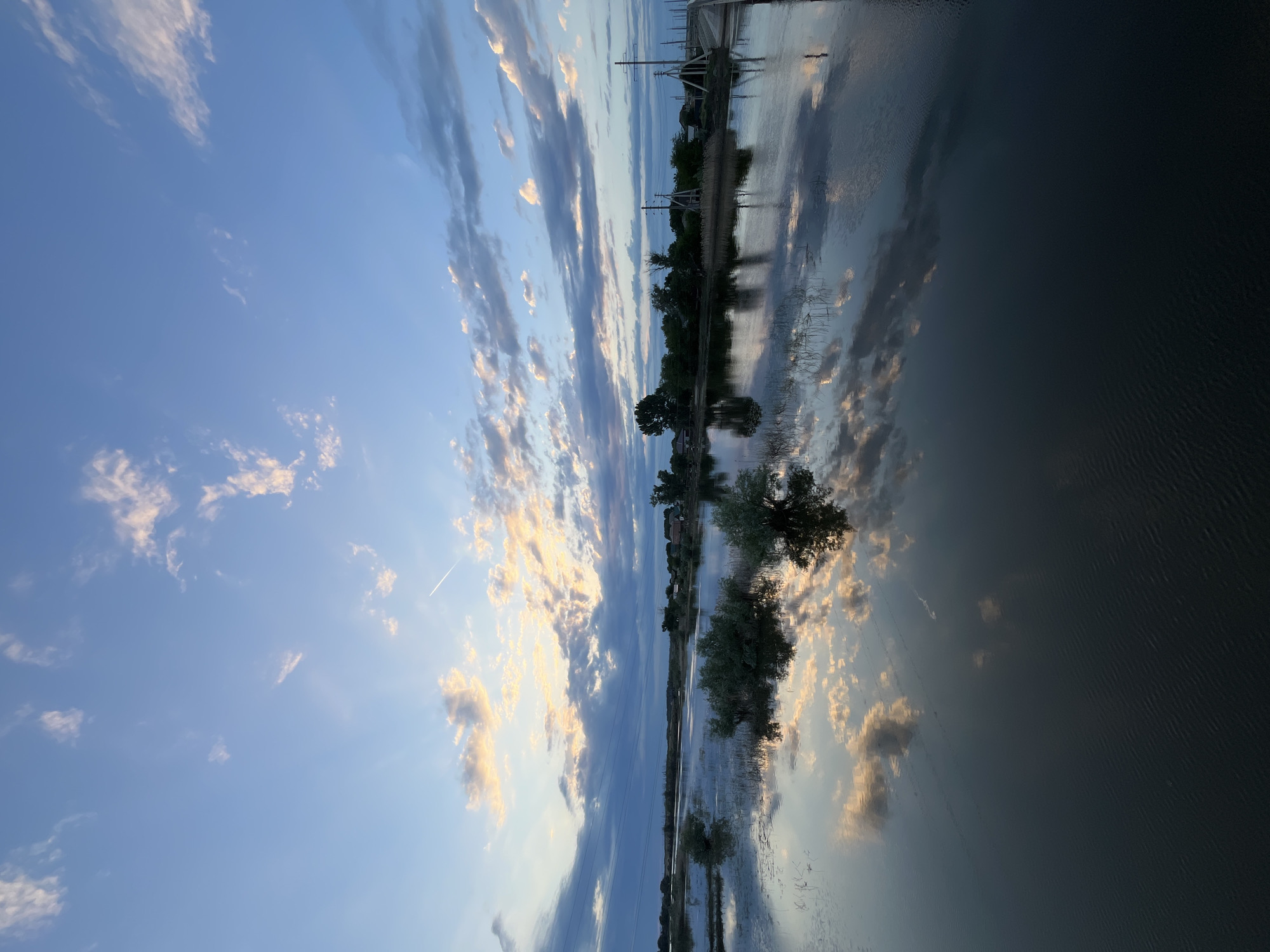 This is a photograph of a calm body of water, likely a river or lake, in the city of Krasnyye Barrikady, Russia. The sky above is a clear blue, with scattered white and pink clouds, suggesting it is either sunrise or sunset. The clouds are reflected in the still water, creating a mirror image. There are several small trees and bushes growing in and around the water. In the distance, there is a treeline with some structures that appear to be houses and other buildings. There are also power lines visible crossing the scene. A bridge or walkway is present on the right side of the image, extending into the water. The water's surface has a slight ripple in the foreground. There are no people or animals visible in the image. The overall scene is peaceful and natural.
