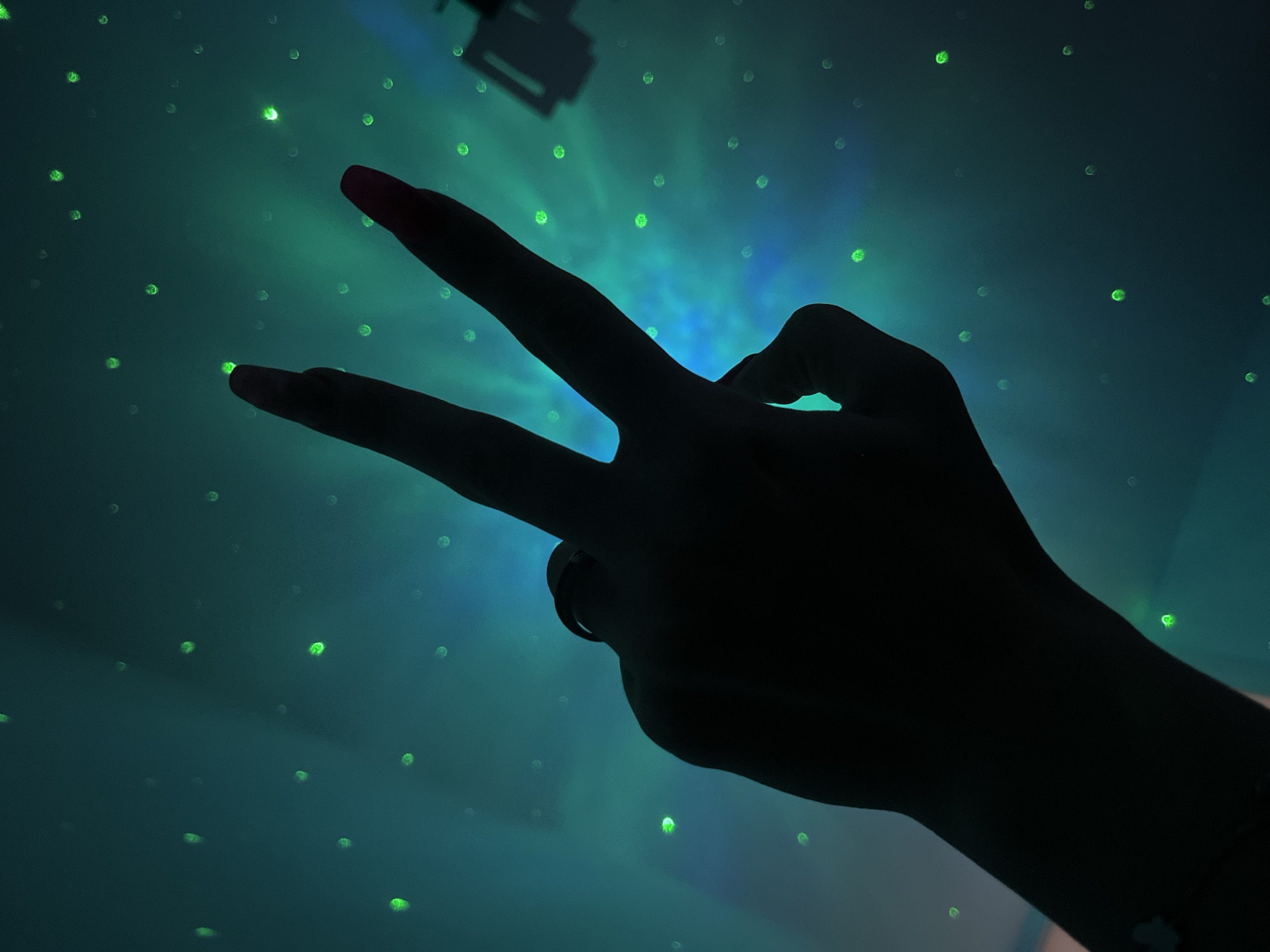 A low-angle shot presents a dark hand making a "peace sign" against a background of blue and green light. The light pattern resembles a night sky with evenly spaced green dots. The person's fingers are silhouetted, but the tips of the index and middle fingers are illuminated with a pinkish-red color. A ring can be seen on one of the fingers. The location cannot be determined from the image, as the background provides insufficient data to identify the city of Plovdiv, Bulgaria.