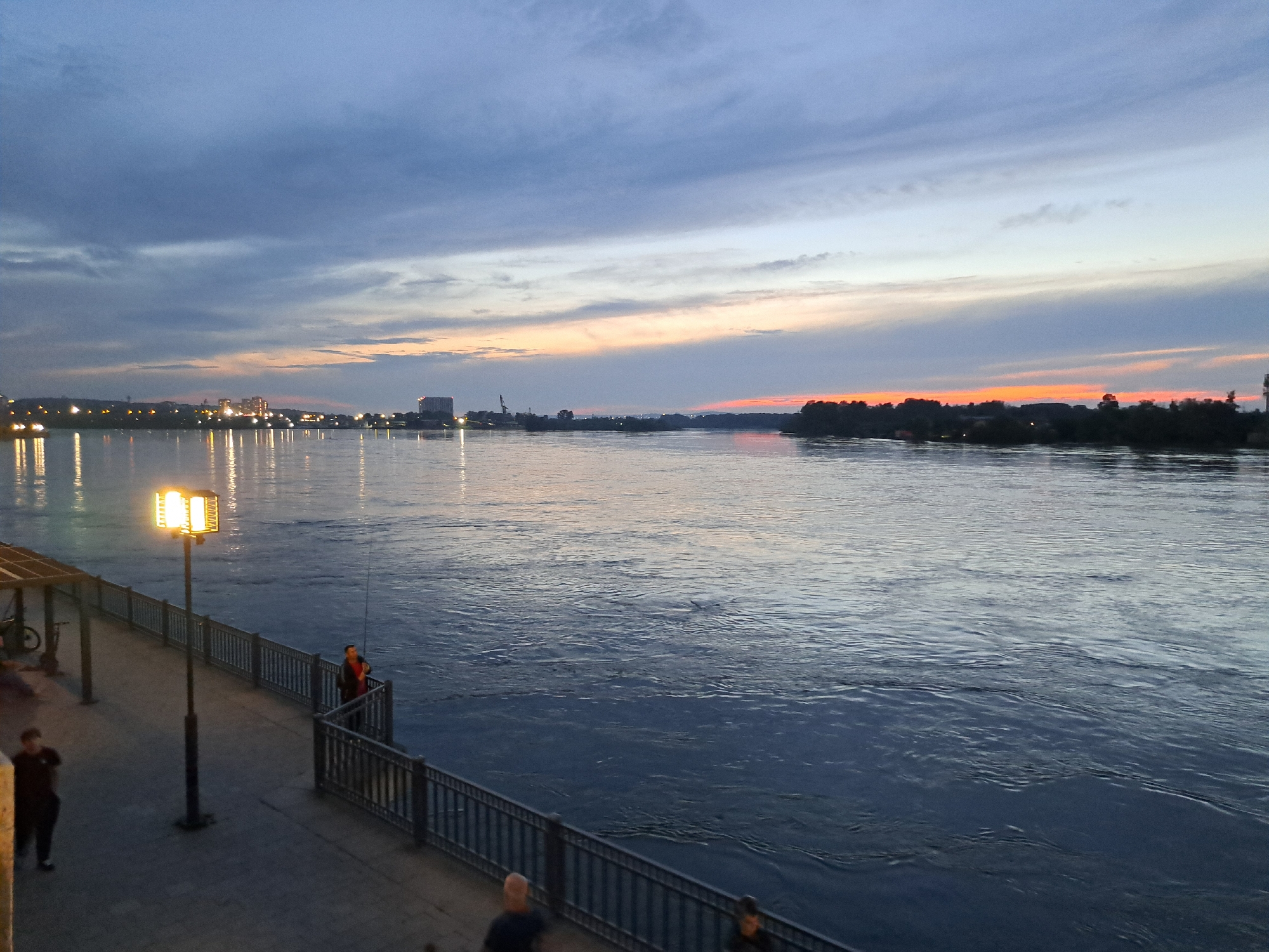 The image depicts a serene evening scene along a riverbank, likely in Irkutsk, Russia, given the context. The sky displays a gradient of twilight colors, with muted blues and grays transitioning into warmer hues of orange and pink near the horizon, where the sun has recently set. Wispy clouds are scattered across the sky, reflecting the fading light.
The foreground features a paved embankment or walkway with a decorative metal railing overlooking a wide, calm river. A solitary streetlamp with multiple illuminated bulbs stands on the embankment, casting a warm glow on the walkway. To the left of the lamp, a portion of an awning or shelter is visible, with a bicycle parked beneath it.
Several individuals are present on the embankment. A person in a dark shirt and pants walks away from the camera on the left side of the walkway. Further along the embankment, near the railing, a person in a red jacket is fishing. Another individual, partially visible, stands near the fishing person. On the opposite side of the river, a distant cityscape is discernible, characterized by scattered lights and silhouettes of buildings and industrial structures, including what appears to be a crane. The river's surface reflects the sky and distant lights, creating a shimmering effect. The overall atmosphere is one of tranquility and the quiet beauty of dusk in an urban riverside setting.