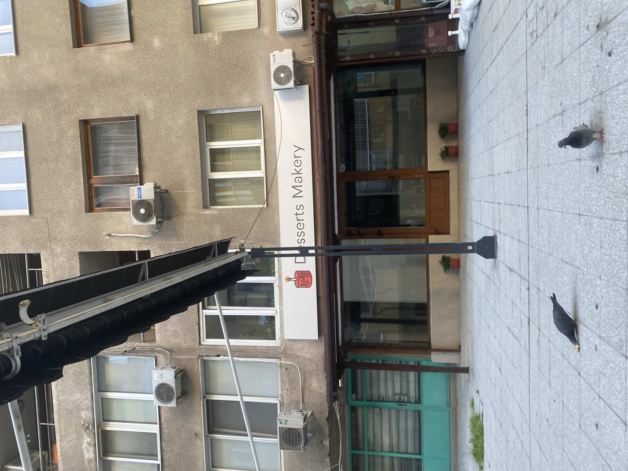The image shows the facade of a building with "Desserts Makery" signage, located in Sofia, Bulgaria. The building has multiple windows and air conditioning units. Two pigeons are on the tiled pavement in front of the building. One is facing towards the right of the image while the other one is facing towards the left.