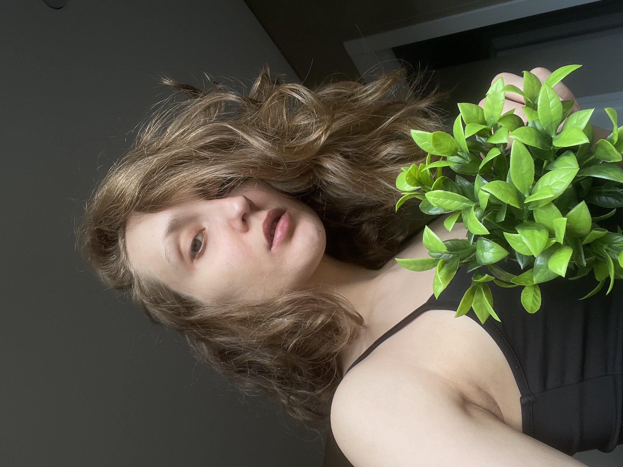 A close-up, rotated portrait features a young individual, likely female, with voluminous, wavy light brown hair. Her face is partially visible, showing one eye, a nose, and slightly parted lips with a neutral to pensive expression. She holds a small artificial plant with vibrant green leaves close to her right shoulder and chest. A black strap from her garment, possibly a tank top or dress, is visible on her shoulder. The background is a plain, dark grey wall, suggesting an indoor setting with soft, diffused lighting. There are no discernible location cues within the image specific to Подшивалово, Russia, nor is any text visible.