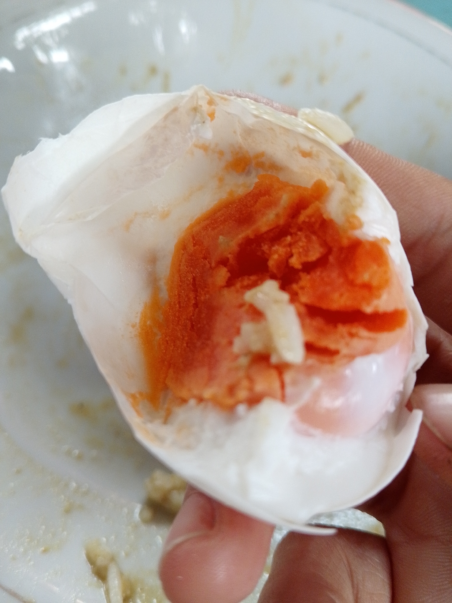 The image shows a close-up of a person's hand holding a peeled salted egg. The egg yolk is a vibrant orange and appears to be slightly crumbly, with some white pieces of egg white visible. The egg white is translucent and forms a shell-like casing around the yolk. The background is blurry, but appears to be a white plate with some food remnants, suggesting a meal setting. The lighting is bright, highlighting the textures and colors of the egg. There are no identifiable landmarks or context to suggest a specific location within Lhokseumawe, Indonesia. The scene is focused solely on the salted egg and the hand holding it.