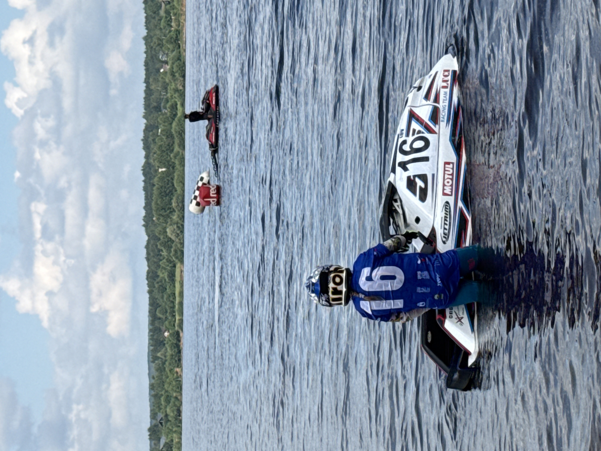 In Perm, Russia, on a body of water with ripples, a person wearing a blue racing suit with the number "16" on the back and a helmet stands in the water next to a white jet ski. The jet ski has "16" printed on its side, along with the words "JETTRIM," "MOTUL," and "RACING TEAM LUCI." In the background, another person is riding a red jet ski, and there are buoys with red and checkered patterns in the water. A tree-lined shore is visible in the distance under a cloudy sky.