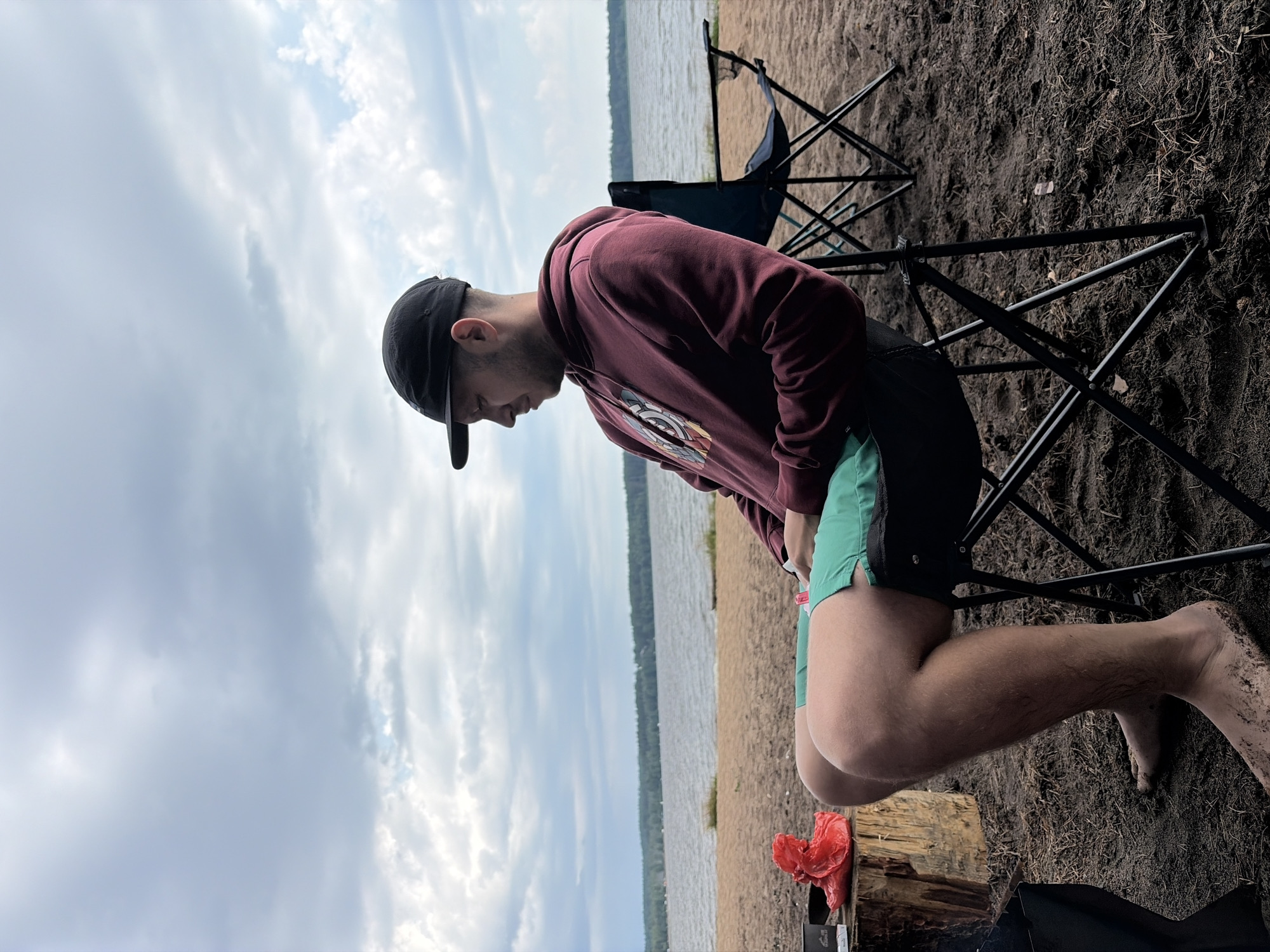 A man wearing a maroon hoodie, green shorts, and a black baseball cap sits in a black folding chair on a sandy beach. He is looking down and to his left. The beach is next to a body of water, with a treeline visible in the distance. The sky is overcast with clouds. To the man's left, there is a wooden stump with a red plastic bag and a small black container on it. Another black folding chair is visible behind him to his right. The scene appears to be in Vaskelovo, Russia.