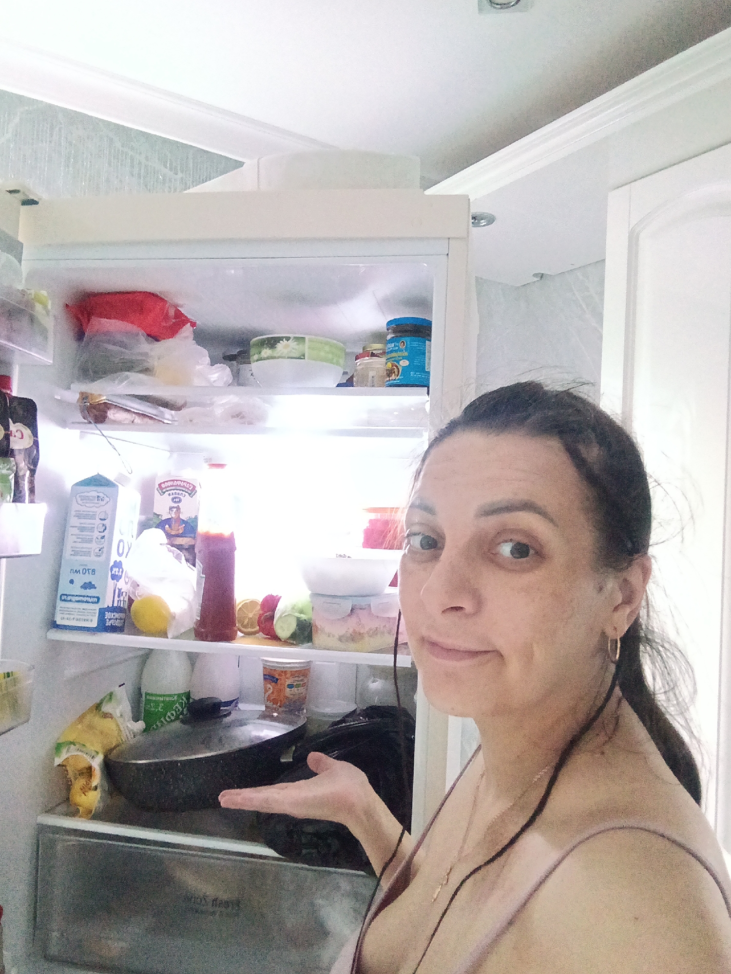A woman stands in front of an open refrigerator in a kitchen. The refrigerator is stocked with various food items, including milk, juice, fruits, and a frying pan. The woman is looking at the camera with a slight smile, and she has her hand extended towards the contents of the refrigerator. The location is Andreyevo, Russia.