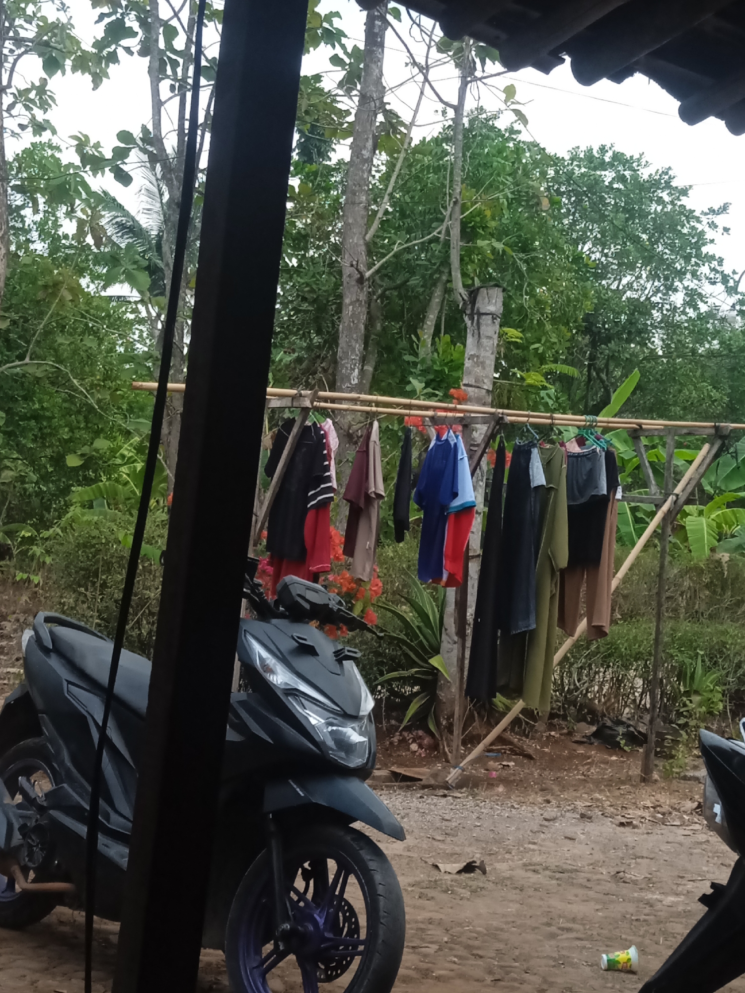 The image depicts an outdoor scene in a rural area, likely Jatisrono, Indonesia. In the foreground, a black motorcycle is partially visible on a dirt and gravel ground. To the right of the motorcycle, a clothesline made of bamboo poles is strung between two wooden supports. Various articles of clothing, including shirts and pants, are hung on the clothesline, some of which appear to be drying. Behind the clothesline, dense green foliage and trees form the background. The sky is overcast. A dark, vertical wooden post is prominently positioned on the left side of the image, obscuring part of the motorcycle and the surrounding environment.