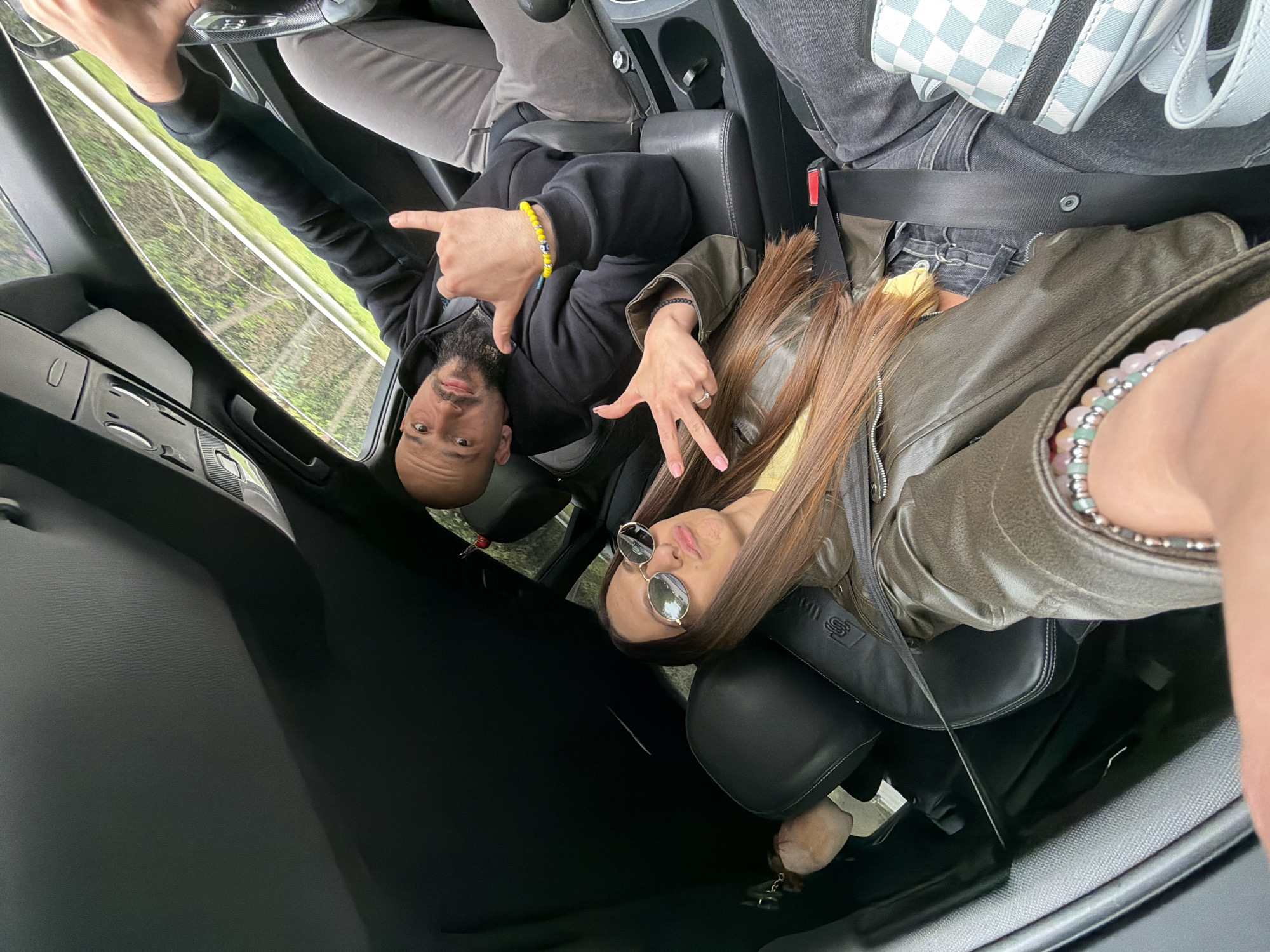A selfie taken from inside a vehicle facing forward depicts two individuals: a bald male in the driver's seat and a female passenger. The male is wearing a black sweatshirt and a yellow bracelet, making a hand gesture with both hands. The female is wearing a yellow top under a green jacket, and sunglasses. She is also wearing bracelets. Both individuals are buckled in with seatbelts. The setting appears to be a car interior, with the road and trees visible through the windshield and sunroof.
There is not enough information to pinpoint the exact location in Varna, Bulgaria.