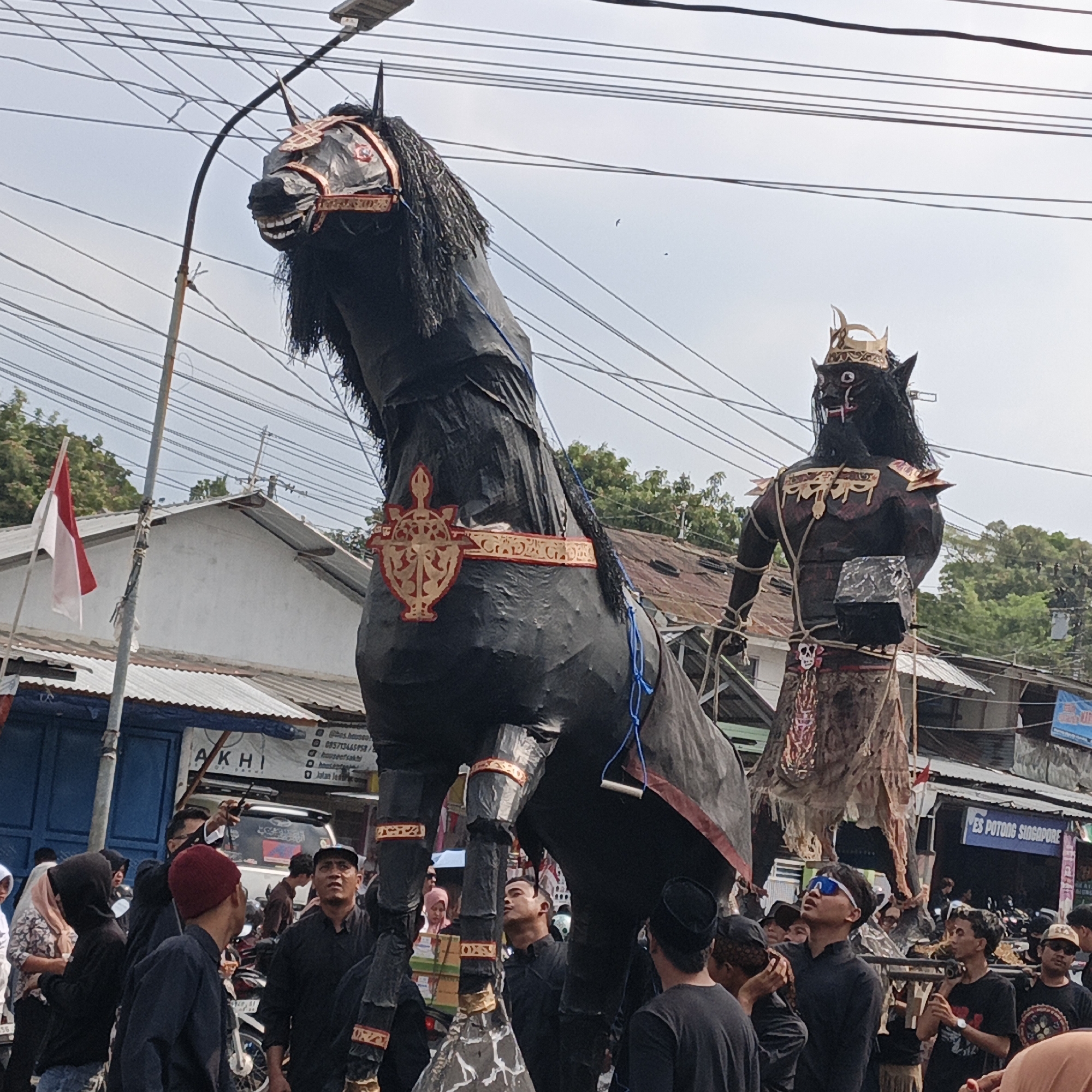 In Randudongkal, Indonesia, a street scene unfolds with a large, black, effigy of a horse dominating the foreground, its head adorned with red and gold decorative elements. Behind the horse, a similarly large effigy of a dark-skinned humanoid figure wearing a golden crown and ornate shoulder pieces is visible. Both effigies appear to be part of a parade or festival. A crowd of people, some dressed in black, are gathered around the effigies, suggesting an event is taking place. Overhead, a network of electrical wires crisscrosses the sky, and a street lamp is mounted on a pole. In the background, buildings with corrugated metal roofs and storefronts are visible, including one with a sign for "ES POTONG SINGAPORE." An Indonesian flag, red and white, flutters on a flagpole. The overall atmosphere suggests a local celebration or cultural event in a semi-urban or rural setting.