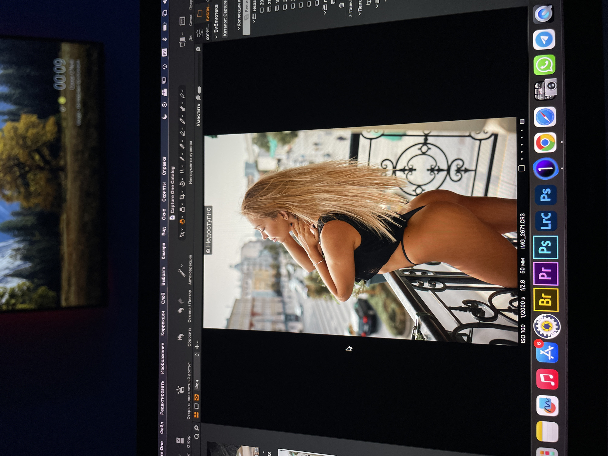 The image depicts a digital display showing a photo being edited in Capture One. The photo features a blonde woman in minimal clothing leaning over a wrought iron balcony. The city skyline is visible in the background. The settings in Capture One show "ISO 100 1/2000 s f/2.8 50 mm IMG_2671.CR3." The desktop also shows a variety of application icons, including Adobe programs and communication apps. The top-left corner shows the time "00:09" on another display. The city is Kazan, Russia.
