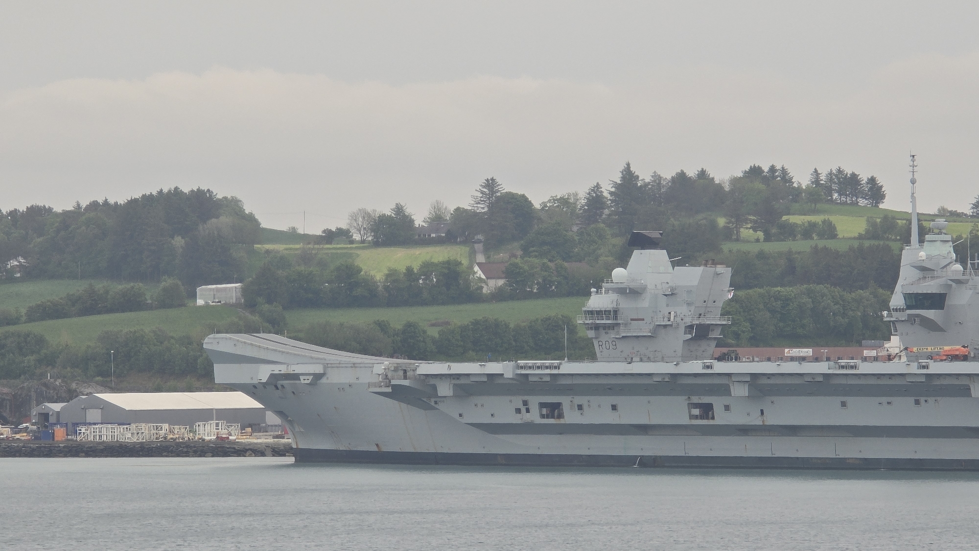A large, grey aircraft carrier, identified by the hull number "R09" prominently displayed on its aft island (superstructure), dominates the central and foreground of the image. The ship exhibits a distinctive 'ski-jump' bow and two island superstructures on its flight deck. On the deck, several objects are visible, including containers, one of which bears the text "Minitrans." Closer to the forward island, an orange vehicle or crane-like structure is present, with the text "EKSTE LIFT.NO" clearly visible on its side.
The aircraft carrier rests in a calm body of water, likely a bay or fjord. The background features a verdant, hilly landscape rising behind the vessel. These hills are densely covered with trees and punctuated by several buildings: smaller residential-style houses nestled among the greenery further up, and larger, more industrial structures, such as a prominent white warehouse, closer to the shoreline.
The scene is set on an overcast day, with a uniformly grey sky above. No specific activity or people are discernible on the ship or the surrounding land. The overall impression is one of a large vessel stationary in a natural, possibly Nordic or Northern European, coastal environment.