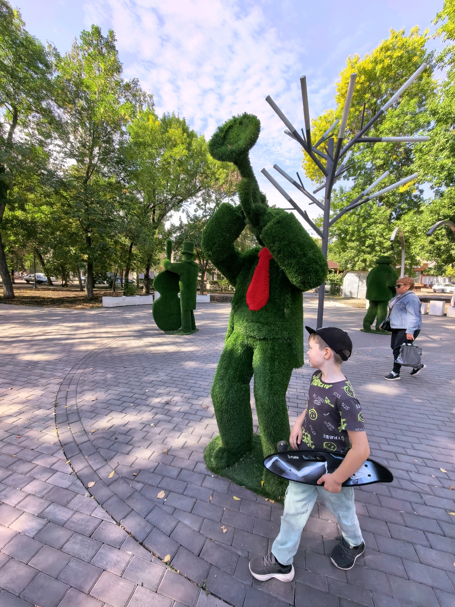 The image depicts a park scene in Sterlitamak, Russia, on a sunny day. In the foreground, a young boy wearing a black baseball cap, a dark t-shirt with neon green lettering, and light blue pants walks on a paved walkway, holding a black skateboard. He is looking to his left, towards a large, anthropomorphic topiary sculpture covered in green material, resembling a figure with its arms raised and a red tie.
Behind the boy and the main topiary figure, there are other green topiary sculptures in the background, including one that appears to be playing a musical instrument and another with a hat. A woman with blonde hair, wearing a denim jacket, dark pants, and carrying a black bag, walks in the mid-ground. The park features mature trees with green and yellow foliage, a paved pathway with a curved pattern, and scattered leaves on the ground. In the distance, there are cars and buildings visible through the trees, suggesting an urban park setting. The sky is blue with scattered white clouds. The overall impression is a vibrant and playful public space.