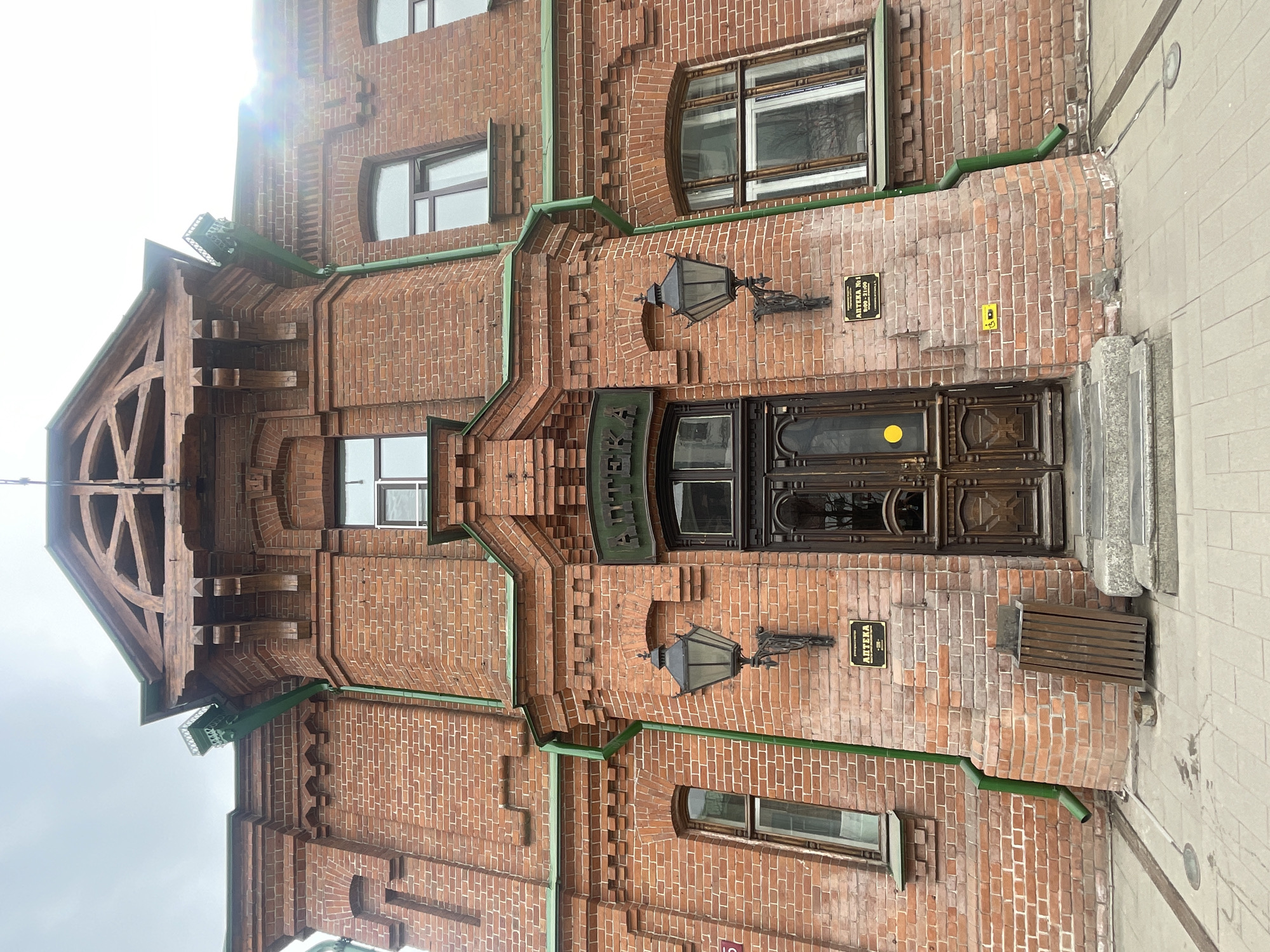 This is a daytime photograph of the facade of an old, ornate brick building in Krasnoyarsk, Russia. The building is constructed predominantly from red and reddish-brown brick, featuring intricate patterns, stepped cornices, and decorative elements characteristic of late 19th or early 20th-century architecture.
The main subject is the building's front, which features a prominent, dark wooden double door with carved panels and glass inserts, serving as the main entrance. Flanking the door are two elaborate, dark metal lanterns mounted on decorative brackets. Above the entrance, a green sign bears the Cyrillic text "АПТЕКА," meaning "Pharmacy."
Further signage is visible on plaques mounted on the brickwork. One clearly reads "АПТЕКА №1" (Pharmacy No. 1) followed by operating hours: "9:00 - 21:00." A similar sign is partially visible on the other side. A small, yellow rectangular object, possibly a doorbell or intercom button, is located to the right of the entrance. To the far right, a small wooden bench or grate is integrated into the wall.
The building has various window styles, including rectangular and arched windows, some with white frames. Green-painted metal drainpipes run vertically and horizontally across the facade, complementing the green sign. The top left features a distinct gabled roofline with visible wooden truss work and intricate detailing.
The scene is an urban setting, with a light-colored paved area visible on the ground level. The time of day is bright, with sunlight casting some glare and lens flare, particularly noticeable in the upper left portion of the image. No people or activities are visible. The architecture and visible Cyrillic text strongly indicate its Russian location.