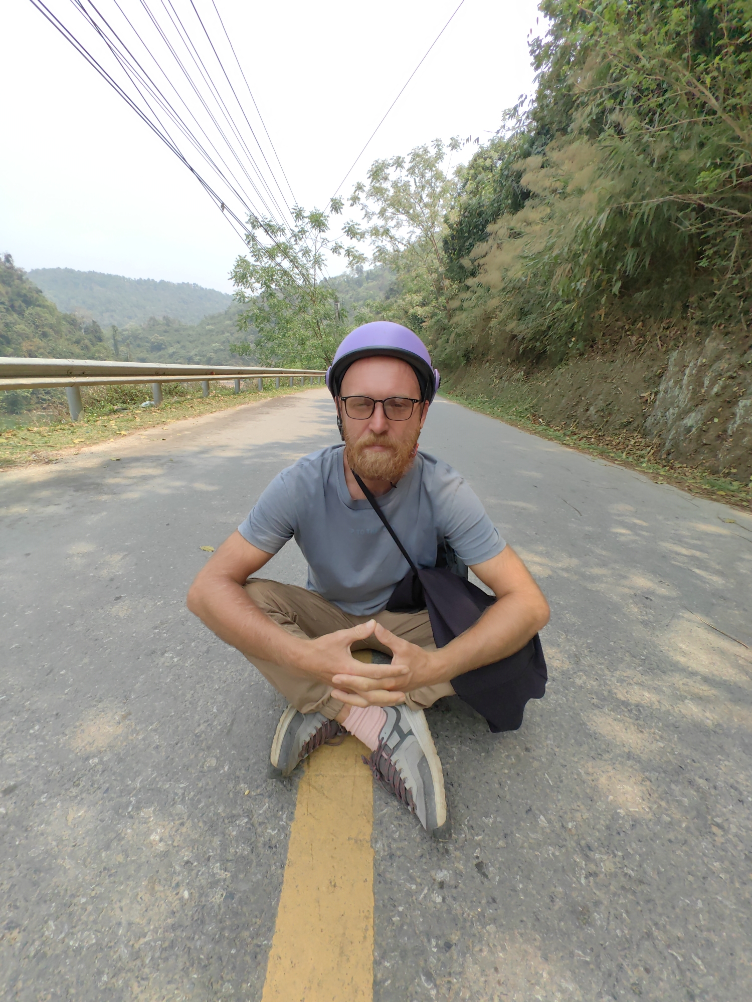 A Caucasian male sits cross-legged on a road in Bảo Lạc, Vietnam. He wears a purple helmet, glasses, and a gray t-shirt with khaki pants. He has a reddish beard. A black bag hangs from his shoulder. His shoes are gray and pink with gray laces. A yellow line runs underneath his legs. The road is gray asphalt. To the left is a silver guardrail and a river. To the right is a green hillside. Overhead are electrical lines and an overcast sky.