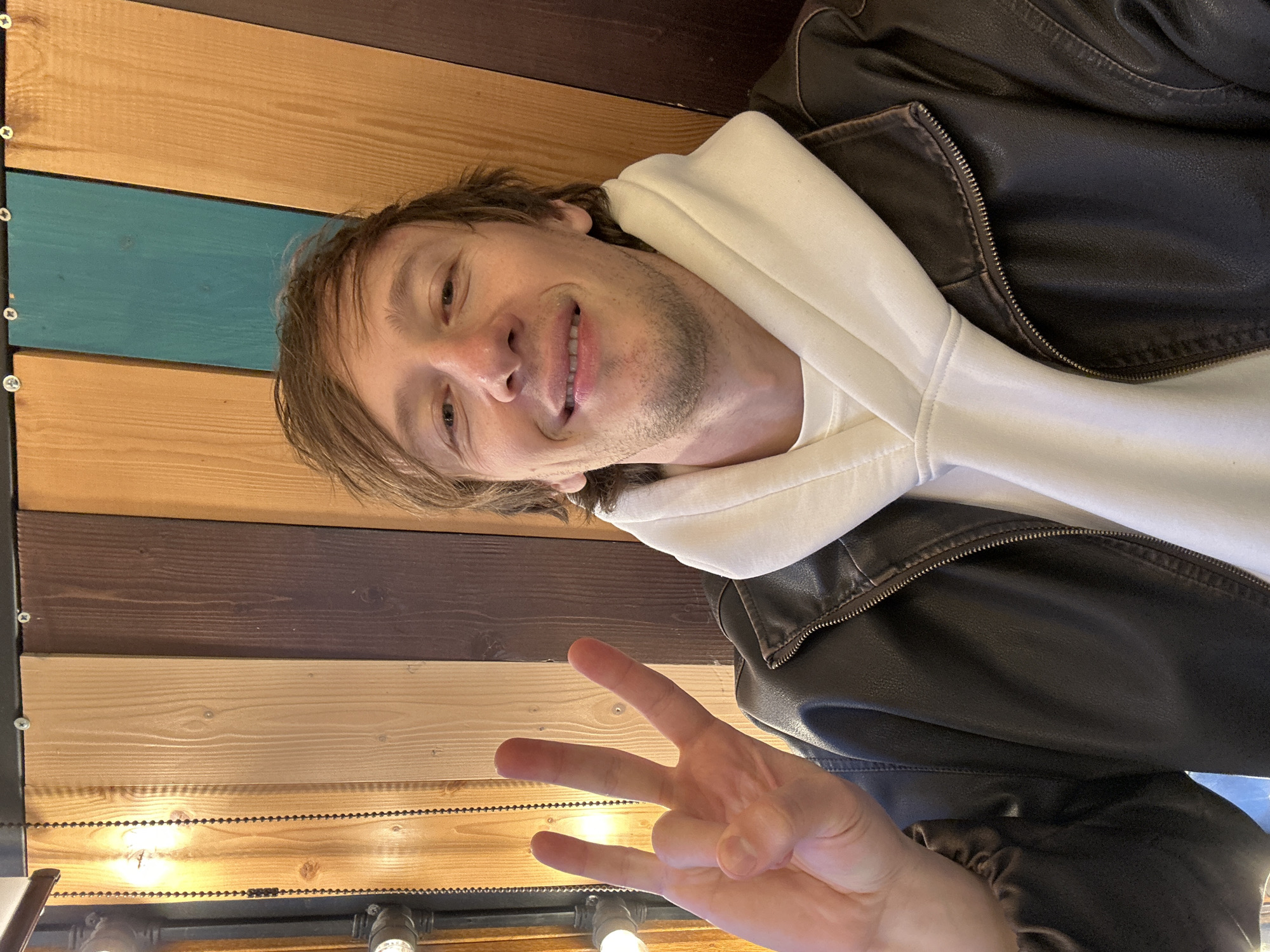 A man is posing for a selfie. He is wearing a brown leather jacket over a white hoodie and is holding up three fingers. The background is a wall made of horizontal wooden planks in various shades of brown and teal. There are lights visible at the bottom of the frame. The man has fair skin and short brown hair. The location is likely a cafe or restaurant in Saint Petersburg, Russia, given the style of the wooden wall and lighting fixtures.