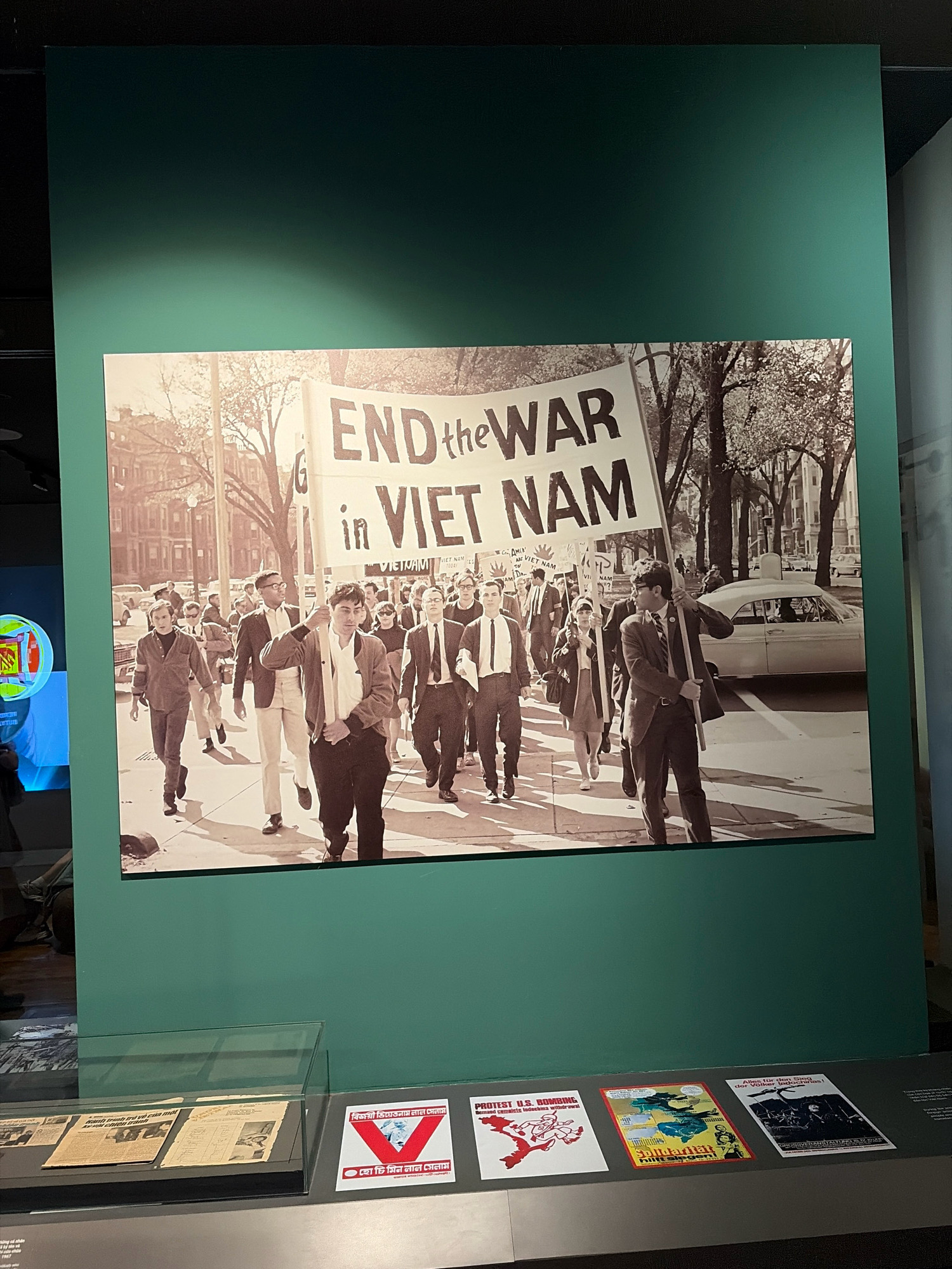 The image displays a historical photograph of a protest, predominantly featuring a large banner that reads "END the WAR in VIET NAM." The photograph is mounted on a dark green wall within a museum exhibit. In the foreground of the exhibit, on a display shelf, are several smaller posters and newspapers. One poster on the right side of the shelf has text in a non-Latin script and a red symbol with a white "V" shape. Another poster to its left has a red graphic of two figures and text that reads "PROTEST U.S. BOMBING Demand complete Indochina withdrawal!". A poster in the center has a collage of images and text, and to its right, a poster with dark imagery and German text. The photograph itself shows a group of people, many appearing to be young adults, marching down a street lined with trees and buildings. Some individuals are carrying smaller signs, and the atmosphere suggests a public demonstration. The presence of a car in the background indicates an urban setting. The image does not provide any visual cues to suggest the protest is located in the city of Đà Nẵng, Vietnam.