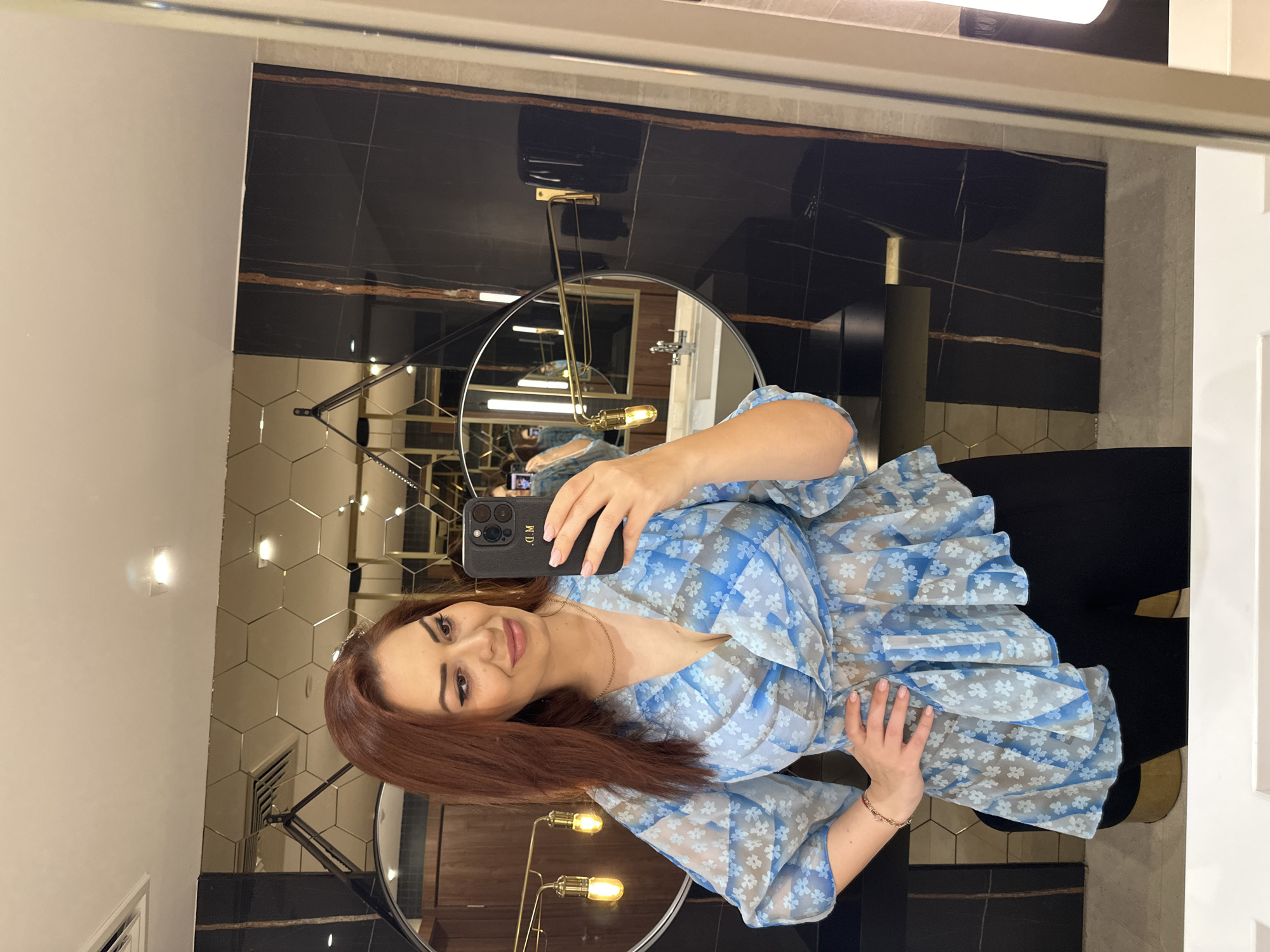 A woman with red hair and wearing a blue floral top is taking a selfie in a bathroom. She is holding a black smartphone with initials "M.D." on it. The bathroom has a circular mirror and modern decor with hexagonal and black tiles, as well as gold fixtures. The location is possibly Kazanlak, Bulgaria, based on the individual's background.