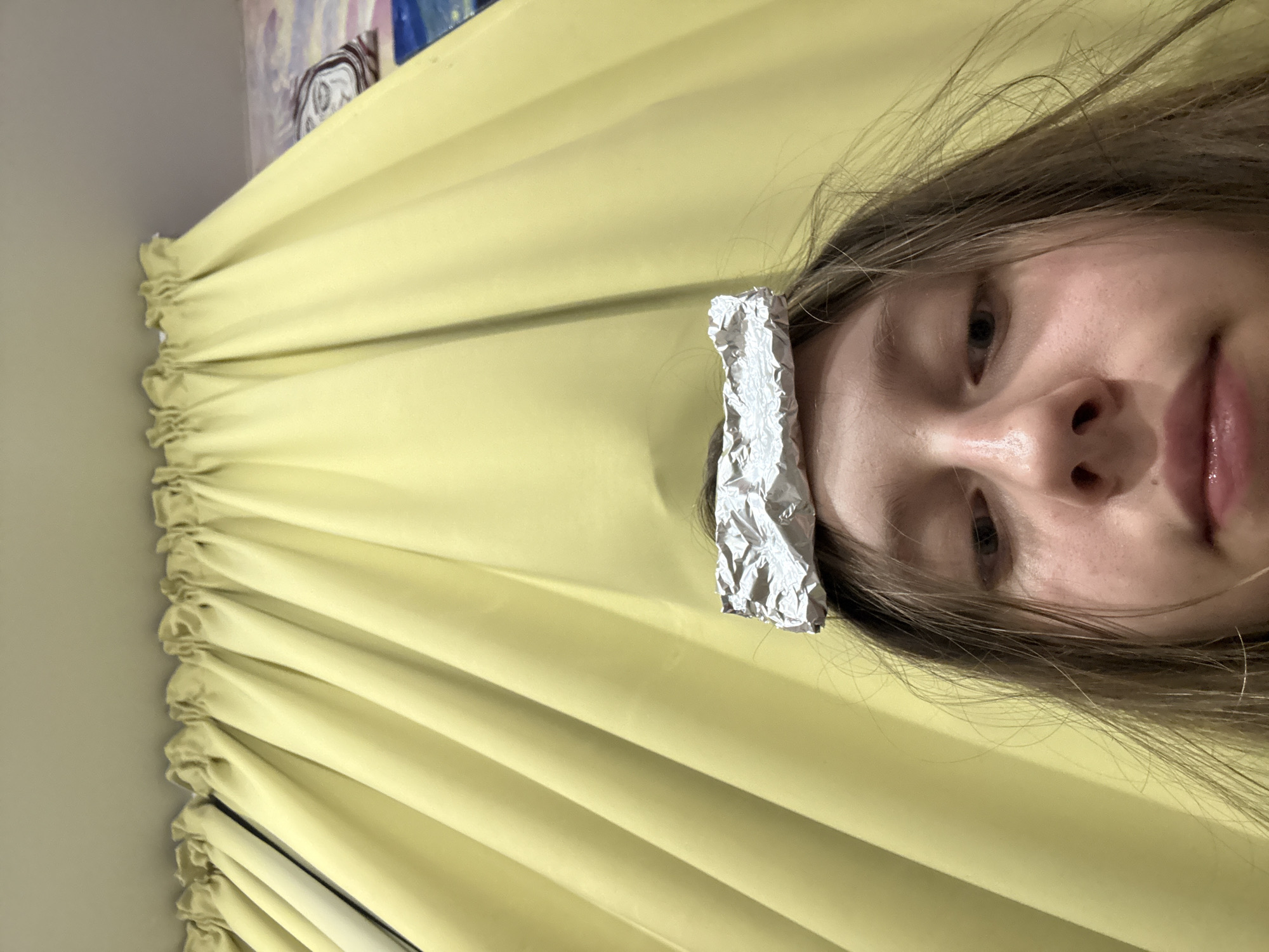 A close-up, low-angle, selfie-style shot shows a young person with light brown hair and fair skin, facing the camera. A crumpled piece of aluminum foil rests on top of their head. The person's eyebrows are furrowed, and their eyes are looking slightly to the left. Their lips are slightly parted. The background consists of yellow curtains, heavily creased and gathered at the top, with a suggestion of a painting or mural visible on the far right. The image appears to be taken indoors. There is no identifiable information suggesting a location in the city of ЖК «Борисоглебское», Russia.
