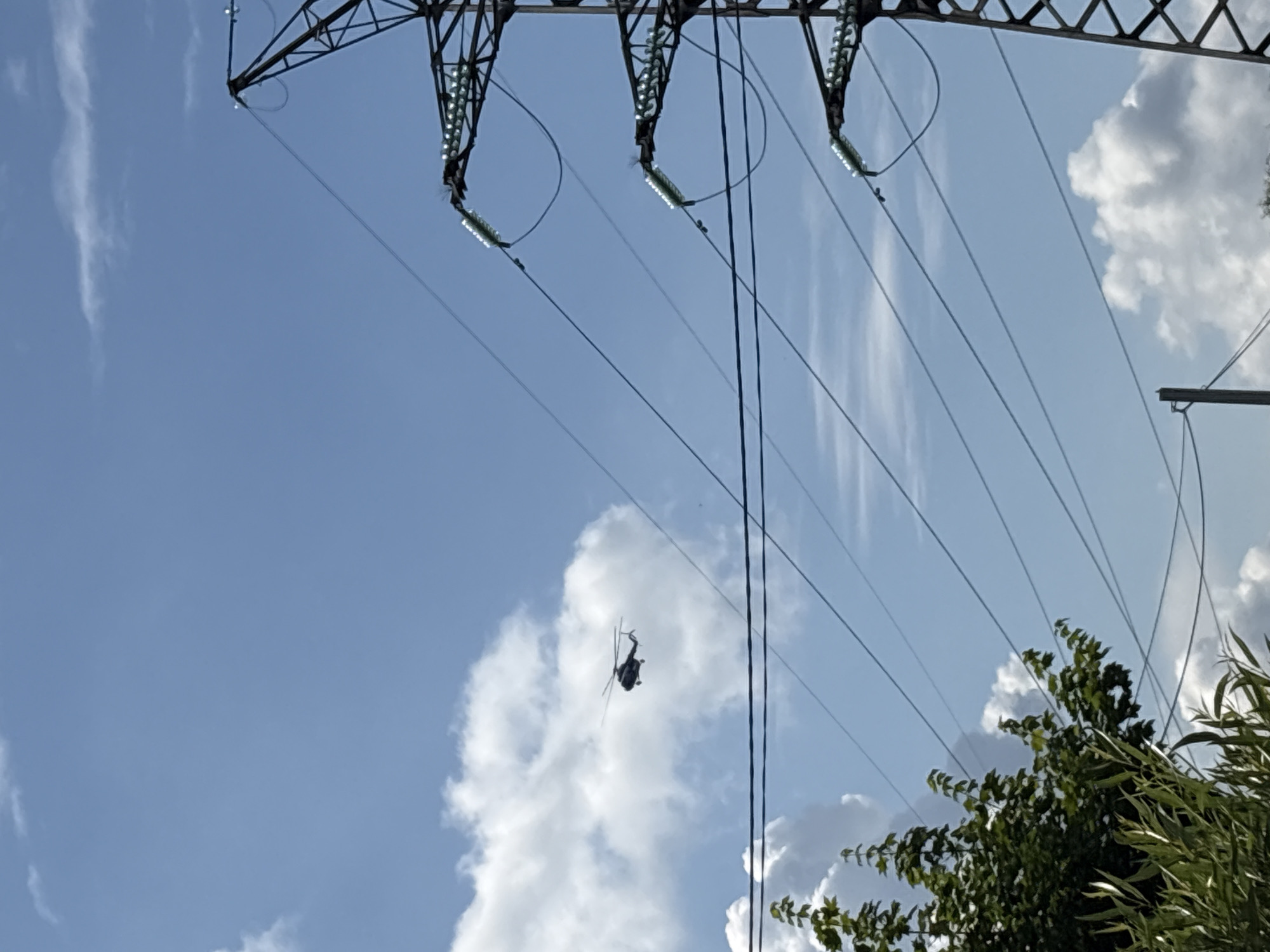 The image shows a dark silhouette of a helicopter flying in a blue sky with scattered white clouds. The helicopter is in the upper left quadrant of the frame. In the right side of the frame, a portion of a tall metal electricity pylon with multiple insulators is visible, supporting several power lines that traverse diagonally across the sky. In the lower left, the tops of green trees and foliage are partially visible. The scene appears to be an outdoor, daytime shot, possibly in or near the city of Chaikovsky, Russia, given the presence of power infrastructure common in such locations. No people or interactions are depicted.