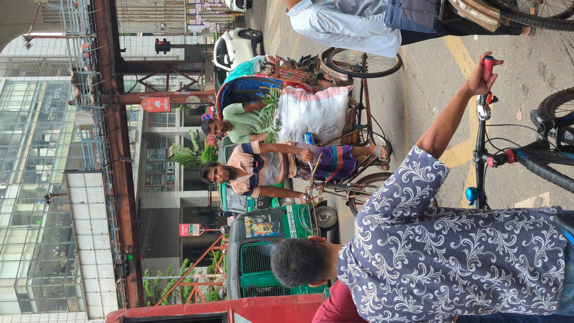 In Dhaka, Bangladesh, a street scene unfolds with a rickshaw carrying two men and a large bag of produce. The rickshaw driver, wearing a striped shirt and a lungi, propels the vehicle forward. Behind him, a passenger in a green t-shirt sits, holding some of the produce. The rickshaw is laden with a white bag filled with green plants. In the background, a green auto-rickshaw is visible, along with parts of a modern building and an elevated structure. A person in the foreground is seen from behind, wearing a patterned shirt and holding the handlebars of a bicycle. Another person in a light blue shirt stands to the right, with a dark bag on their shoulder. The street is paved with yellow lines indicating traffic lanes.