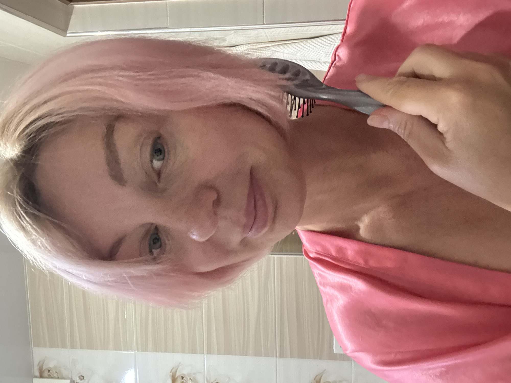 The media file shows a close-up, vertical, selfie-style shot of a woman with light pink hair. She is wearing a pink satin robe. She is holding a gray hairbrush and is in the process of brushing her hair. Her face is turned towards the camera, and her expression appears neutral or slightly amused. The background suggests an indoor setting, likely a bathroom, with tiled walls visible. The lighting is bright, indicating it might be daytime. The woman's makeup is subtle, with noticeable eyebrows and light lip color.