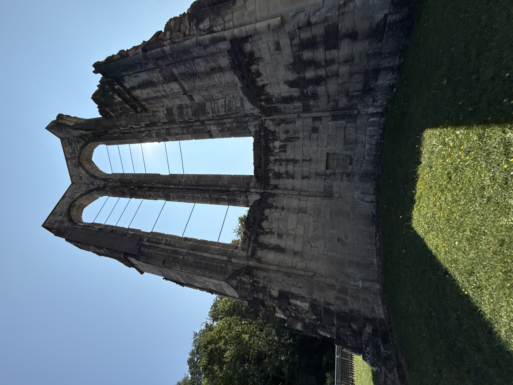 The image shows the ruins of the Walkenried Abbey in Walkenried, Germany. The ruins are made of large stone blocks and appear to be ancient. Two large arched windows are visible, with thin metal bars stretching across them. The sky is clear and blue, and the sun is shining brightly through the windows, creating a lens flare effect. The ruins are surrounded by green grass, and trees can be seen in the background. A fence is also visible to the left of the ruins. The image is taken from a low angle, looking up at the ruins. The overall impression is one of grandeur and decay.