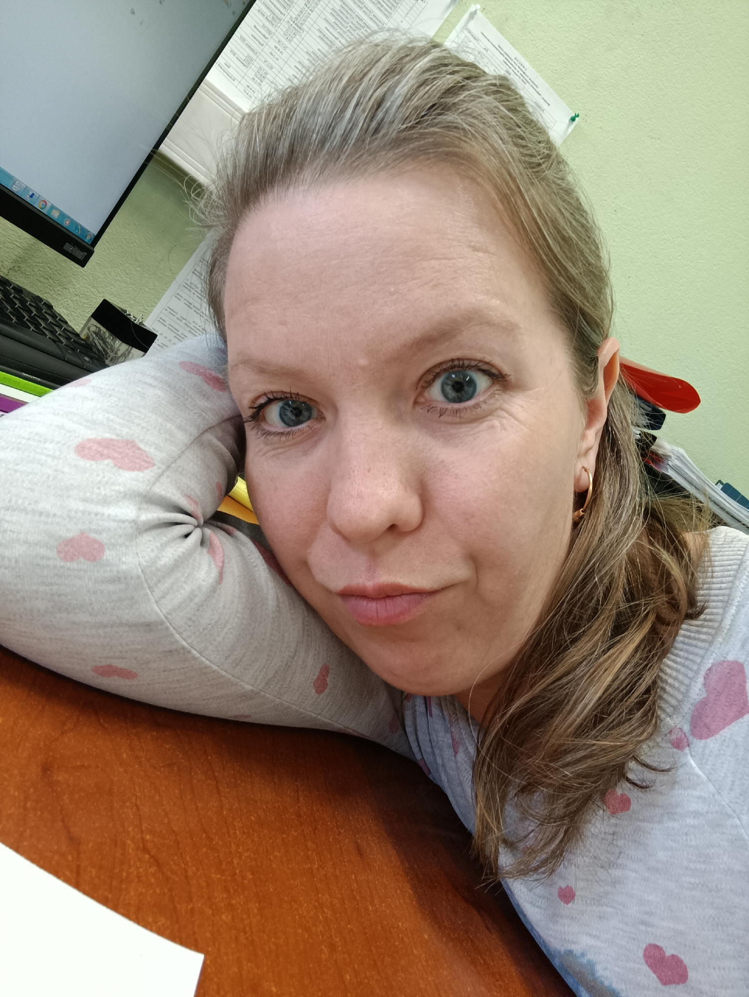 A woman with blue eyes, fair skin, and light brown hair is resting her head on her left arm, which is on a brown wooden surface, likely a desk. She is wearing a light gray sweater with pink heart patterns. A computer monitor and keyboard are visible in the background, along with stacks of papers. The wall behind her is light green. The scene is indoors, likely an office setting. The location is Chaikovsky, Russia.