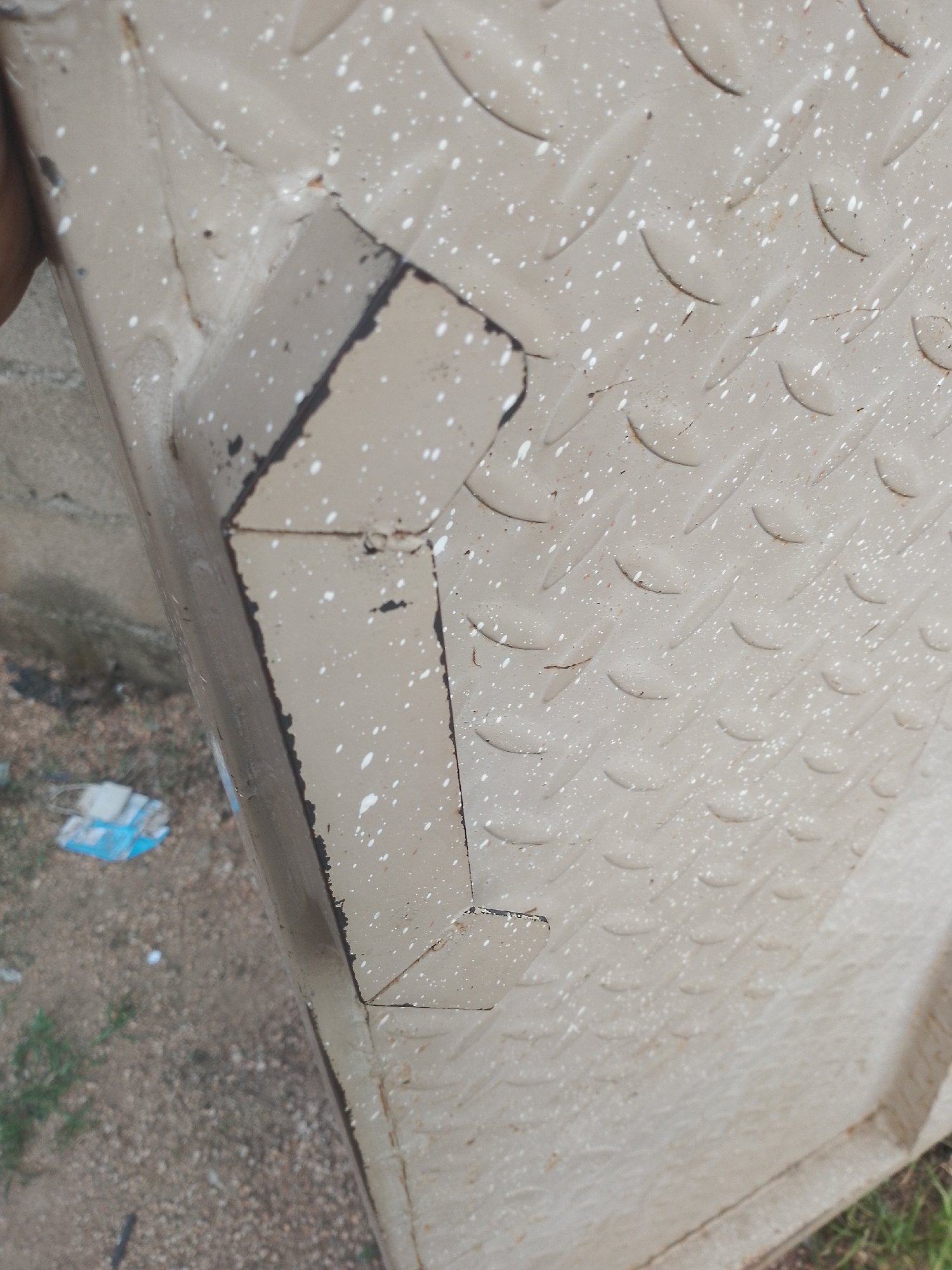 A close-up, angled shot shows a portion of a tan-colored metal surface with a diamond plate texture, speckled with small white paint marks and some dark debris. The metal appears to be part of a larger object, possibly a container or barrier, with folded edges and a seam visible. The background is out of focus, showing a mixture of gravel, dirt, and sparse green vegetation. A blurred, light blue and white object, possibly plastic packaging, rests on the ground to the left. The scene is outdoors, in what appears to be a somewhat unkempt area, suggesting it is not in the city center of Jauro Yinu, Nigeria, but rather in an outlying or industrial section.